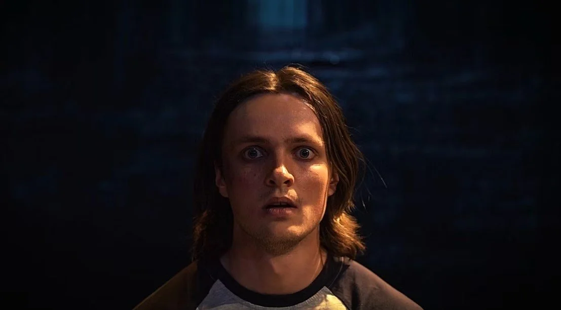 A young man with long hair and a surprised or shocked expression, wearing a black and gray shirt, against a dark background.