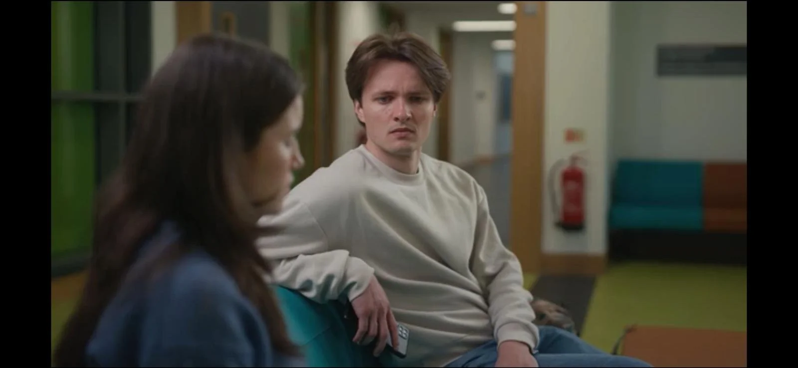 A man and a woman sitting on chairs in a waiting area, facing each other. The man is looking at the woman with a serious or concerned expression, while the woman is looking down. The background shows a hallway with a fire extinguisher and some furnit
