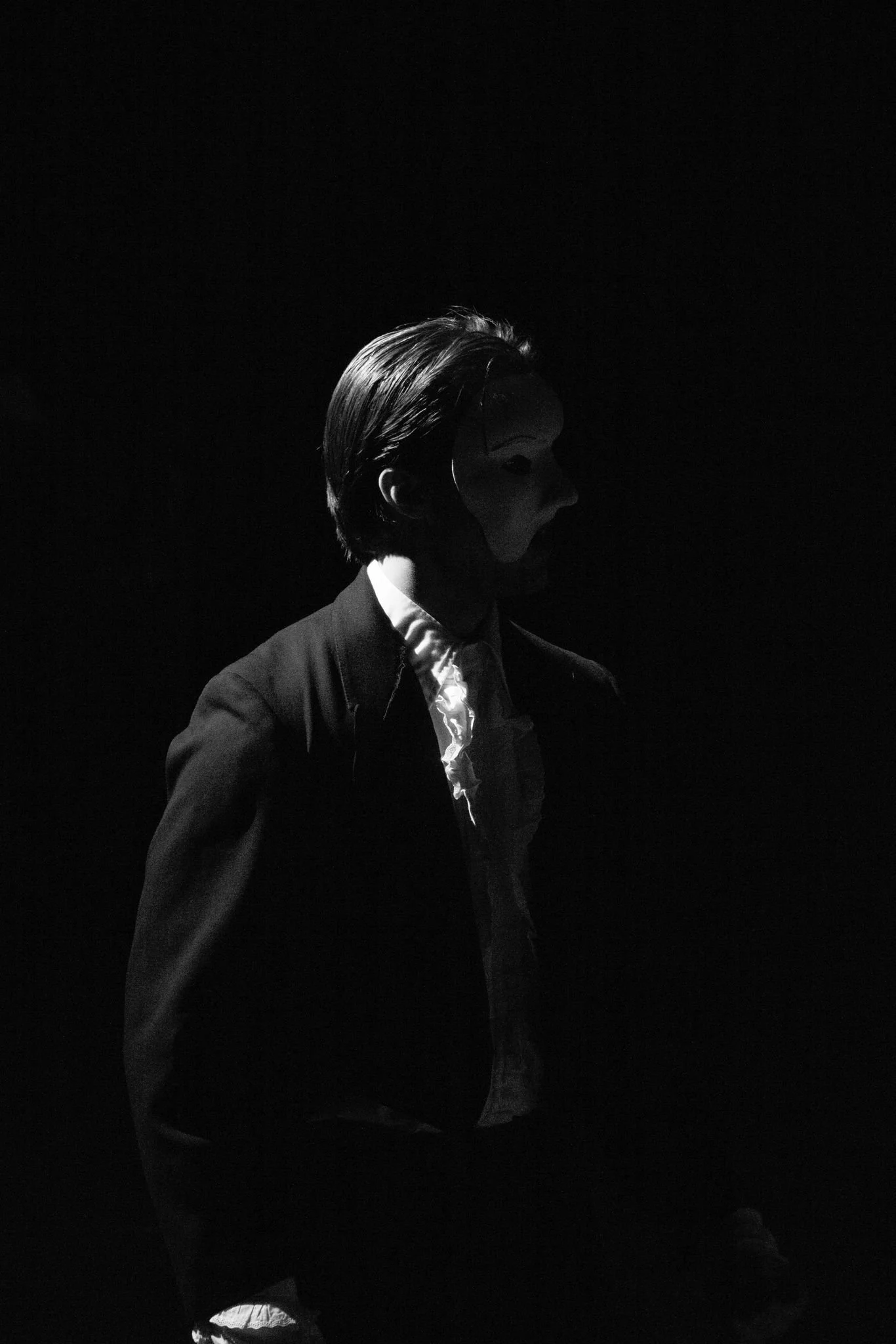 A person with slicked-back hair wearing a mask and dark clothing, standing in a dark environment.