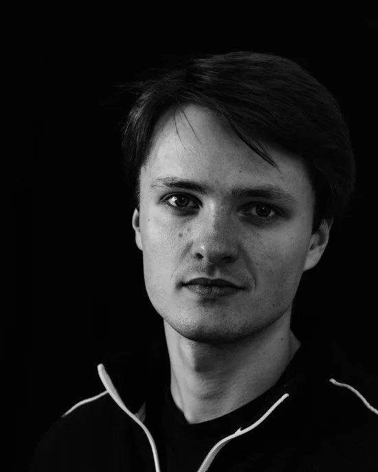 Black and white portrait of a young man with short hair, wearing a zippered jacket, against a dark background.