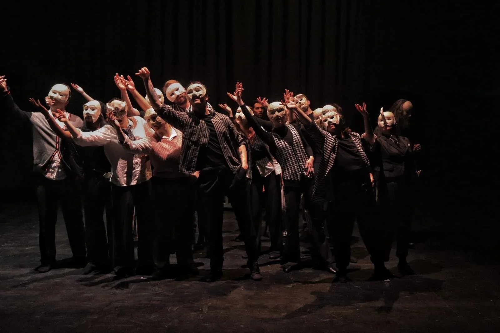 A group of people on stage wearing white masks and dark clothing, performing in a theatrical production with dark background and stage lighting.
