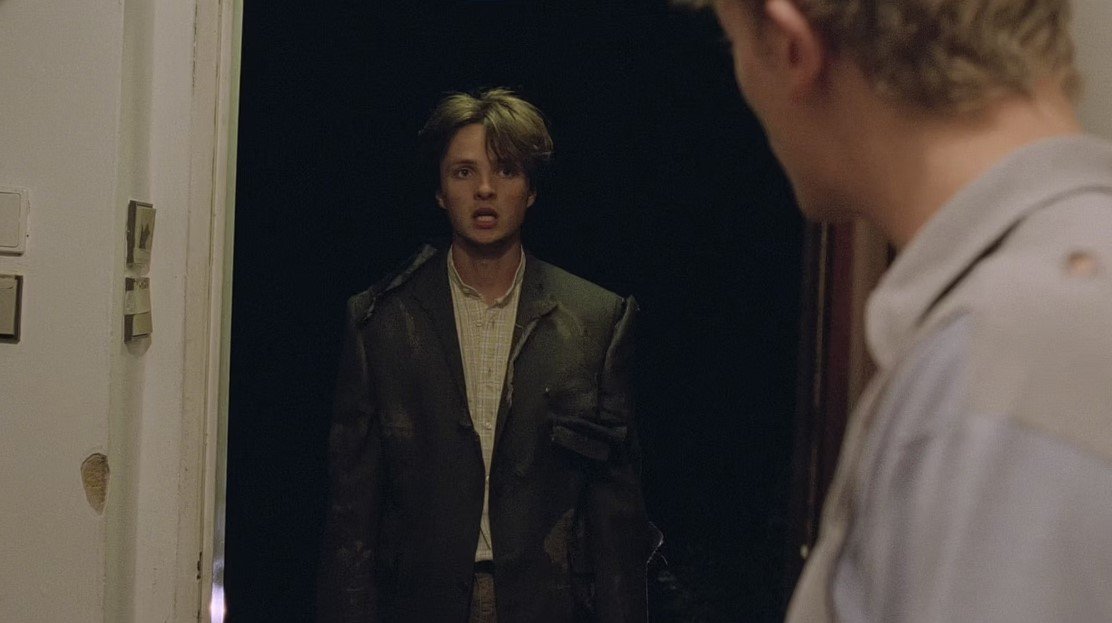 A young boy with messy hair and a distressed expression stands in a doorway, facing a person whose face is partially visible.