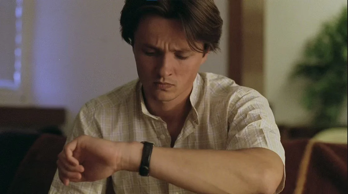 A young man with brown hair looking at his wristwatch with a concerned expression.