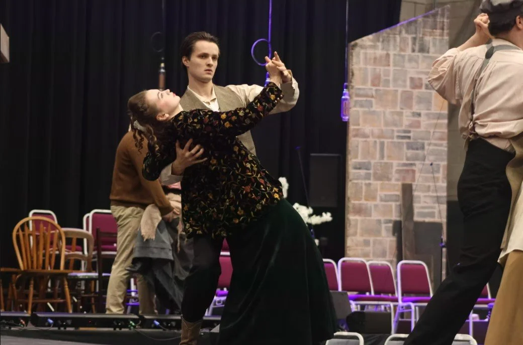 Actors rehearsing on stage with circus rings, with chairs and a brick wall in the background.