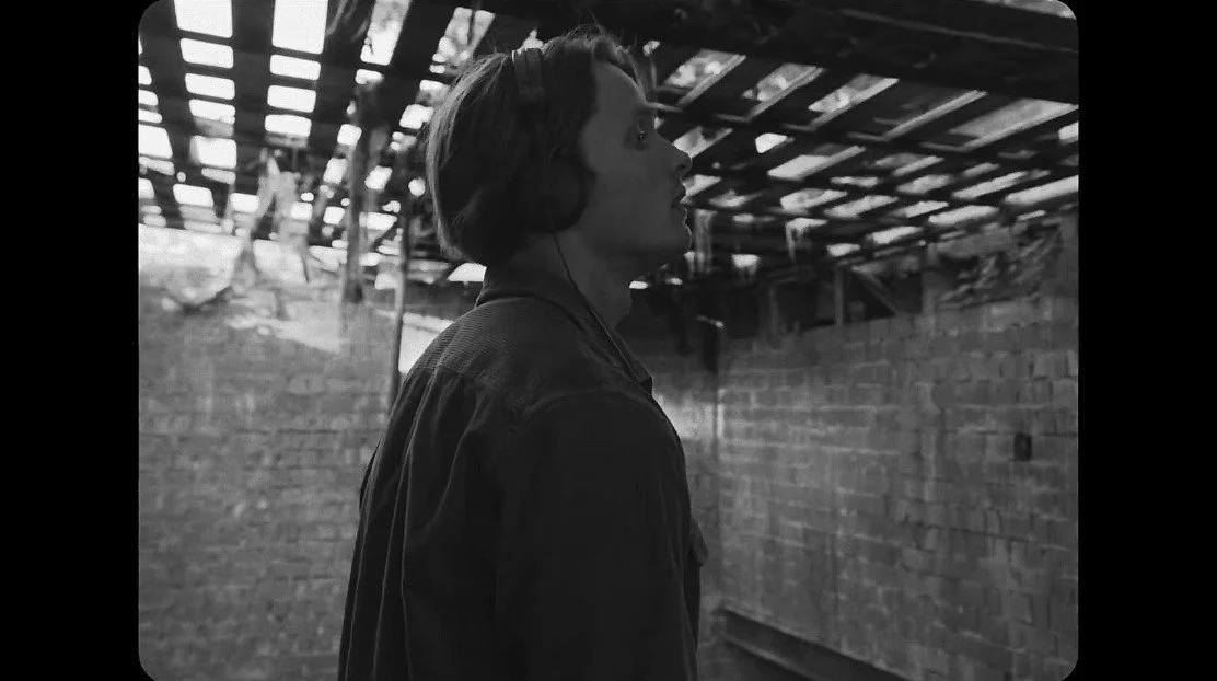 A woman with headphones wearing a jacket, standing under a damaged roof with exposed wooden beams and brick wall, looking to the side in profile, in black and white.