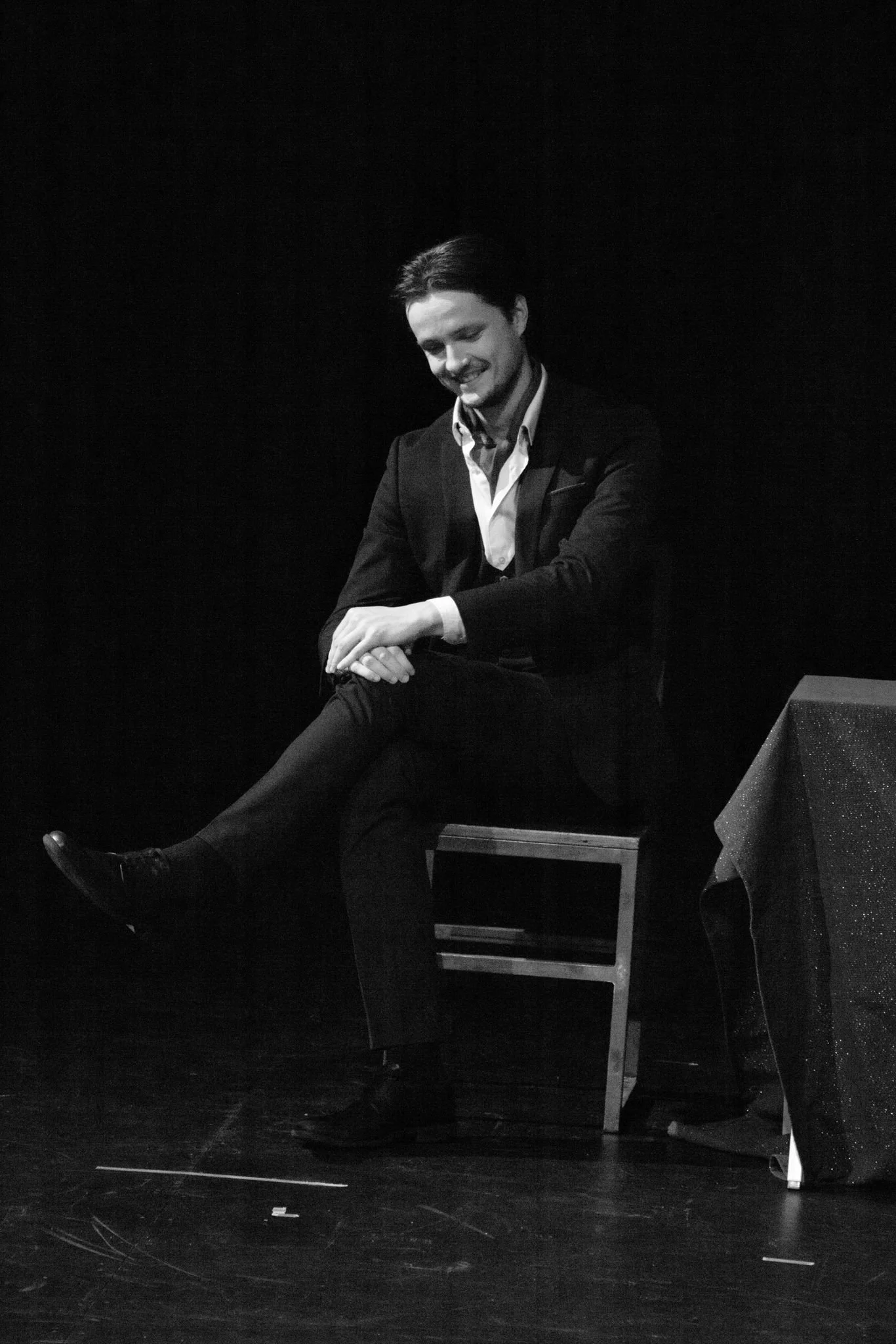 Black and white photo of a young man in a suit sitting with one leg crossed over the other, smiling while looking down, in a dark environment with a table and chairs nearby.