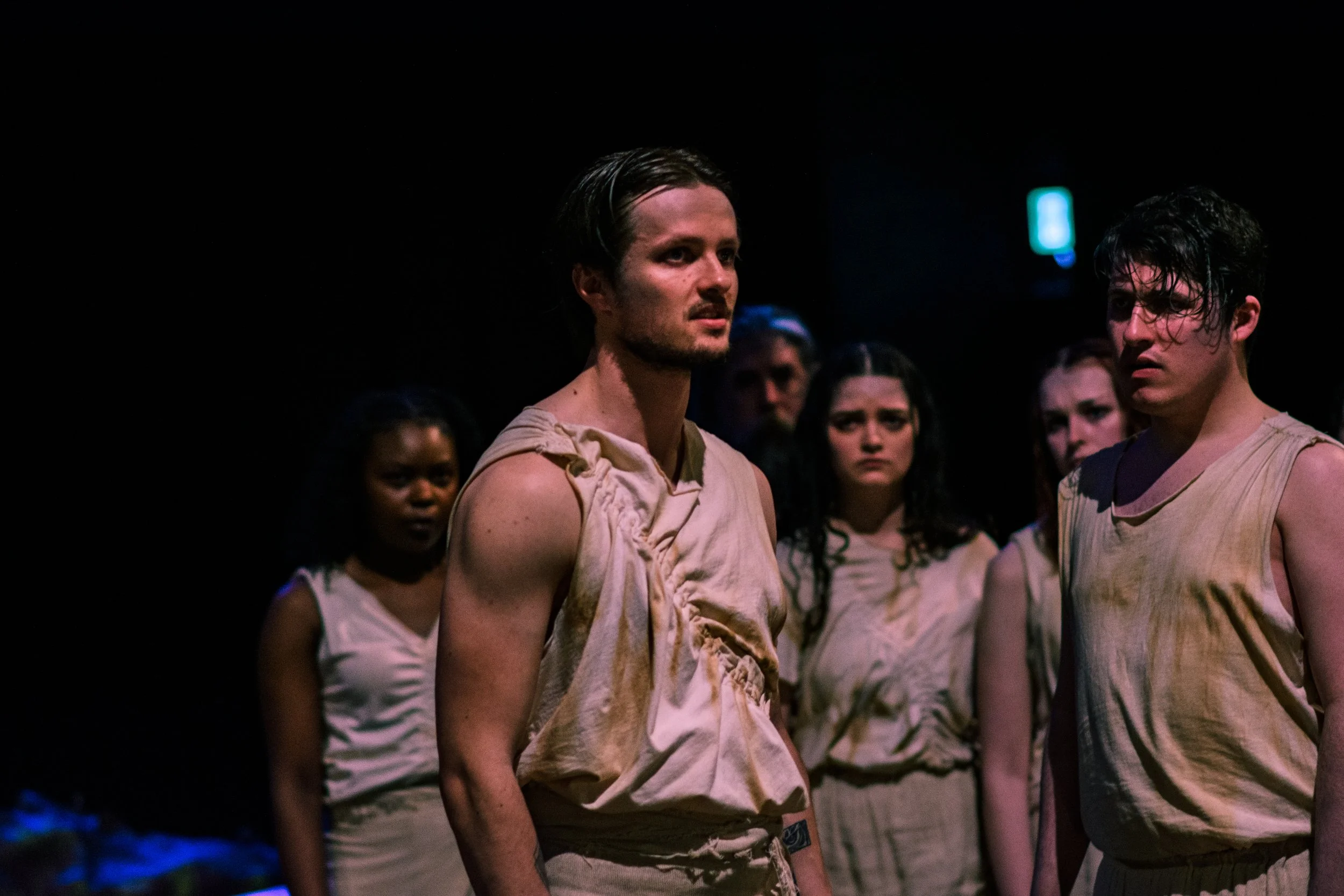 Group of young people with distressed and serious expressions, wearing worn and dirty clothing, standing in a dark environment.