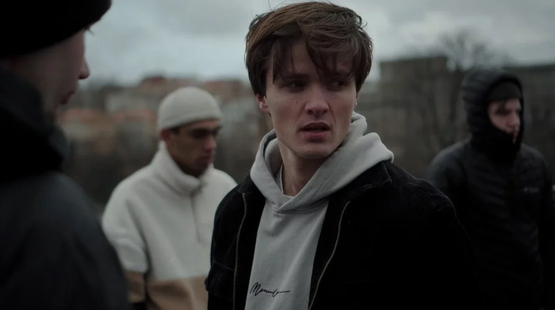 A young man with brown hair wearing a gray hoodie and black jacket looks to his left outdoors with three other people in the background on a cloudy day.