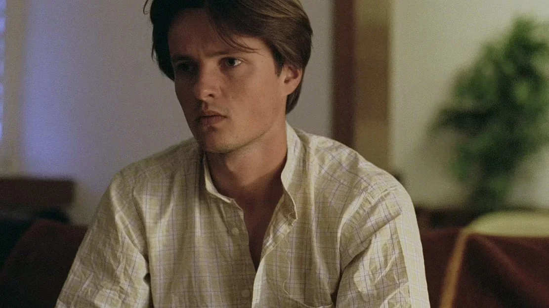 A young man with brown hair wearing a light-colored, checkered shirt sitting indoors with a potted plant in the background.