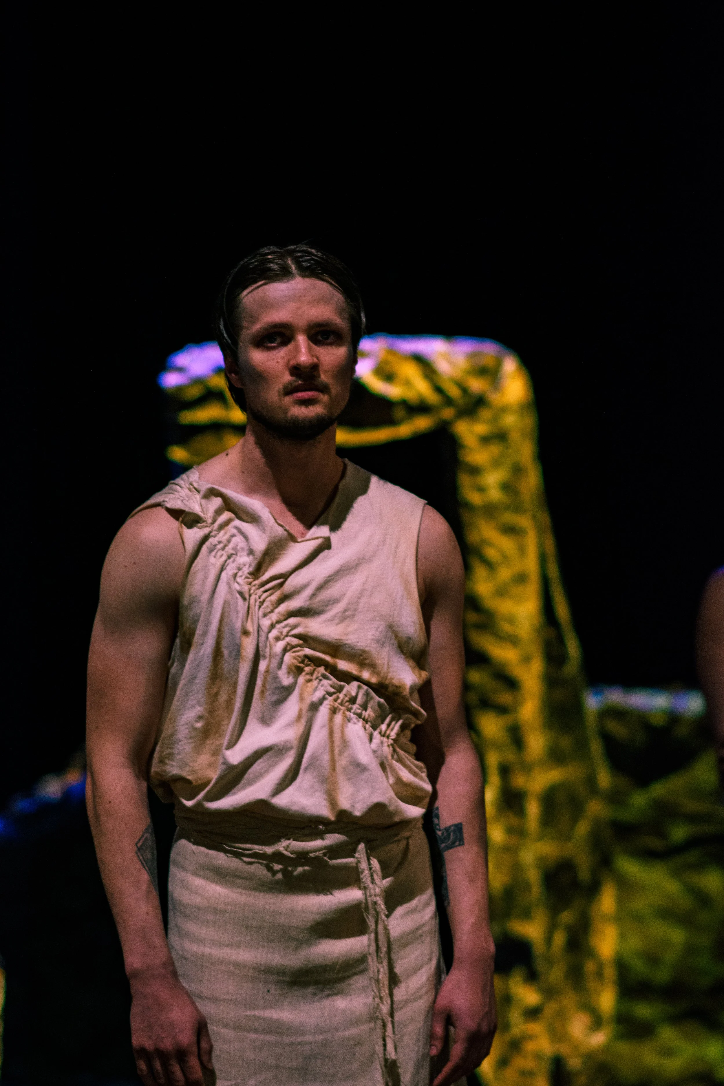 A young man with wet hair and a serious expression stands in front of a dark background with colorful lights. He's wearing a beige, sleeveless, gathered top and matching drawstring pants. Part of another person in yellow camouflage pants is visible b