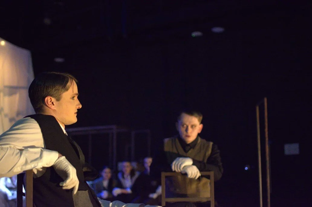 Two young men dressed in vintage attire engaged in a theatrical performance, with one sitting on a wooden chair and the other standing, against a dark background with an audience watching.