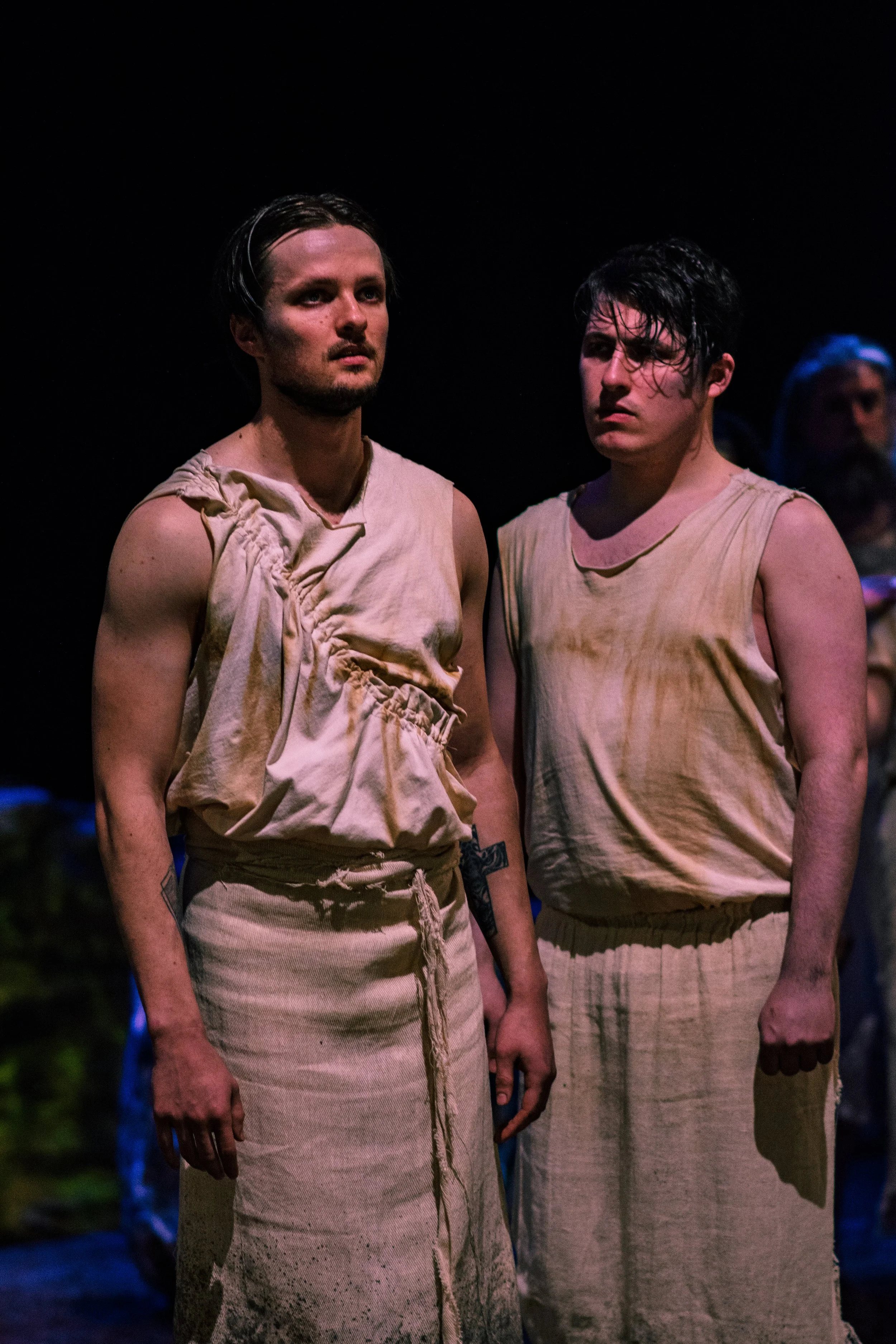 Two women with short dark hair wear beige sleeveless dresses in a dark setting, holding hands, with another person visible in the background.