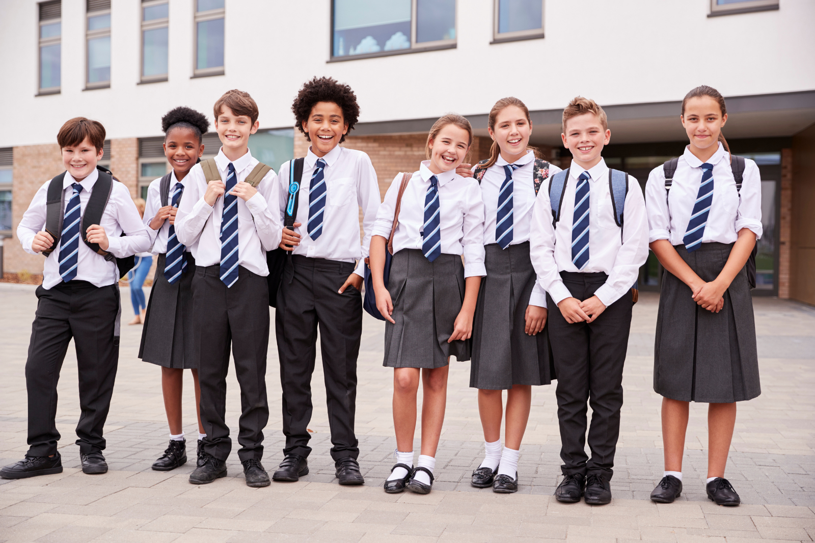 Group of eight diverse school children in uniform standing outside school building, smiling and carrying backpacks.