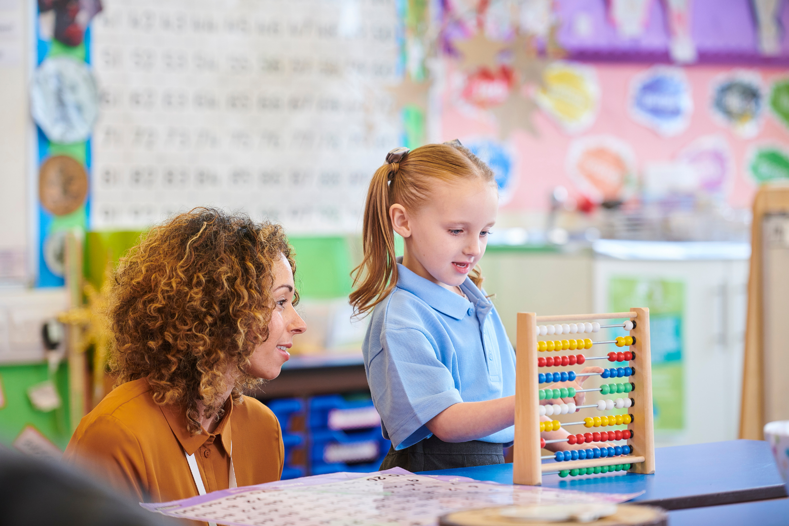 A young girl in a blue school uniform using an abacus during a classroom learning activity, with a teacher observing nearby.