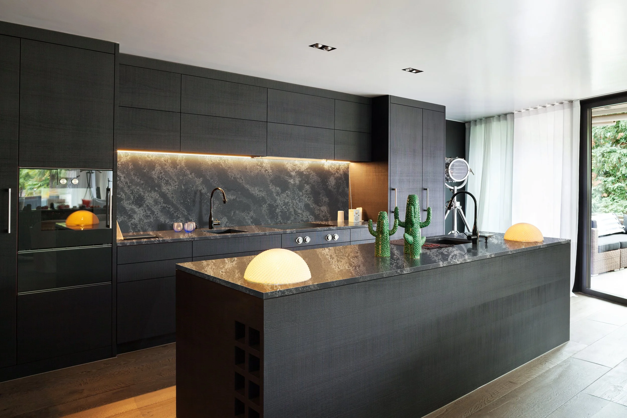 Modern kitchen with dark cabinets, a black marble countertop, and decorative cacti on the island. Large windows with white curtains allow natural light to fill the space.
