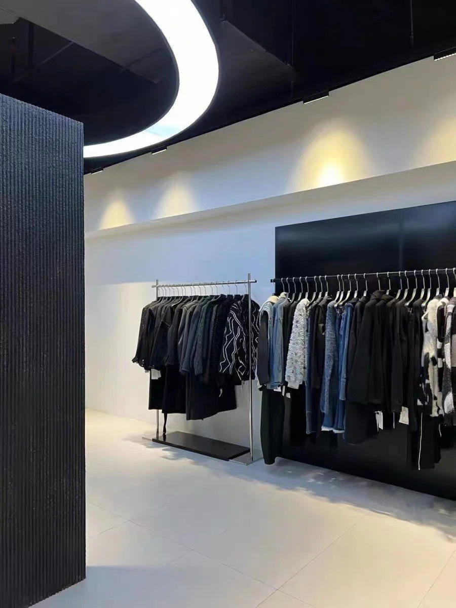 Clothing store display with black and patterned garments on racks against a modern, minimalist interior with white walls, black ceiling, and circular lighting.