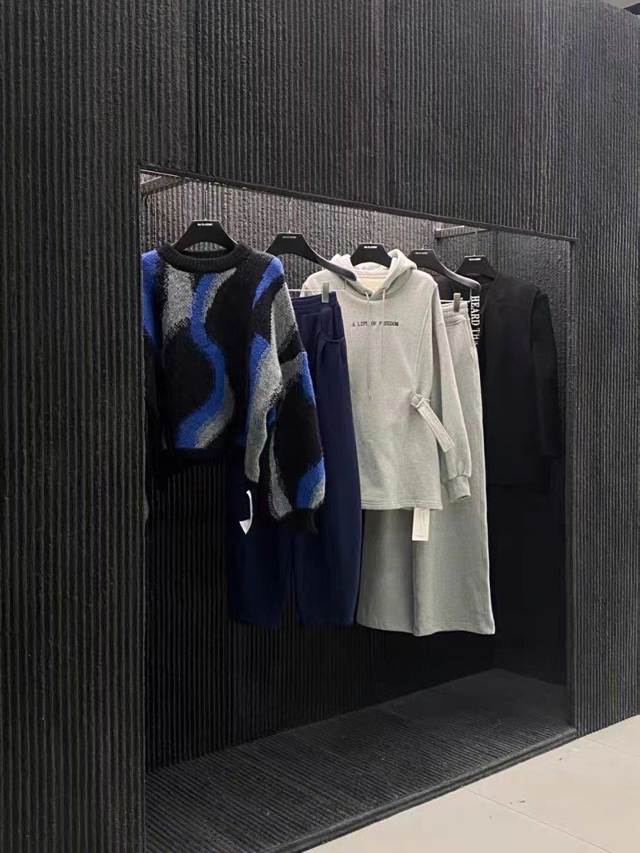 Clothing rack with four displayed outfits, including a black, blue, gray, and white patterned sweater, navy blue pants, a white hoodie with text, a white skirt, and a black jacket, against a black textured wall.