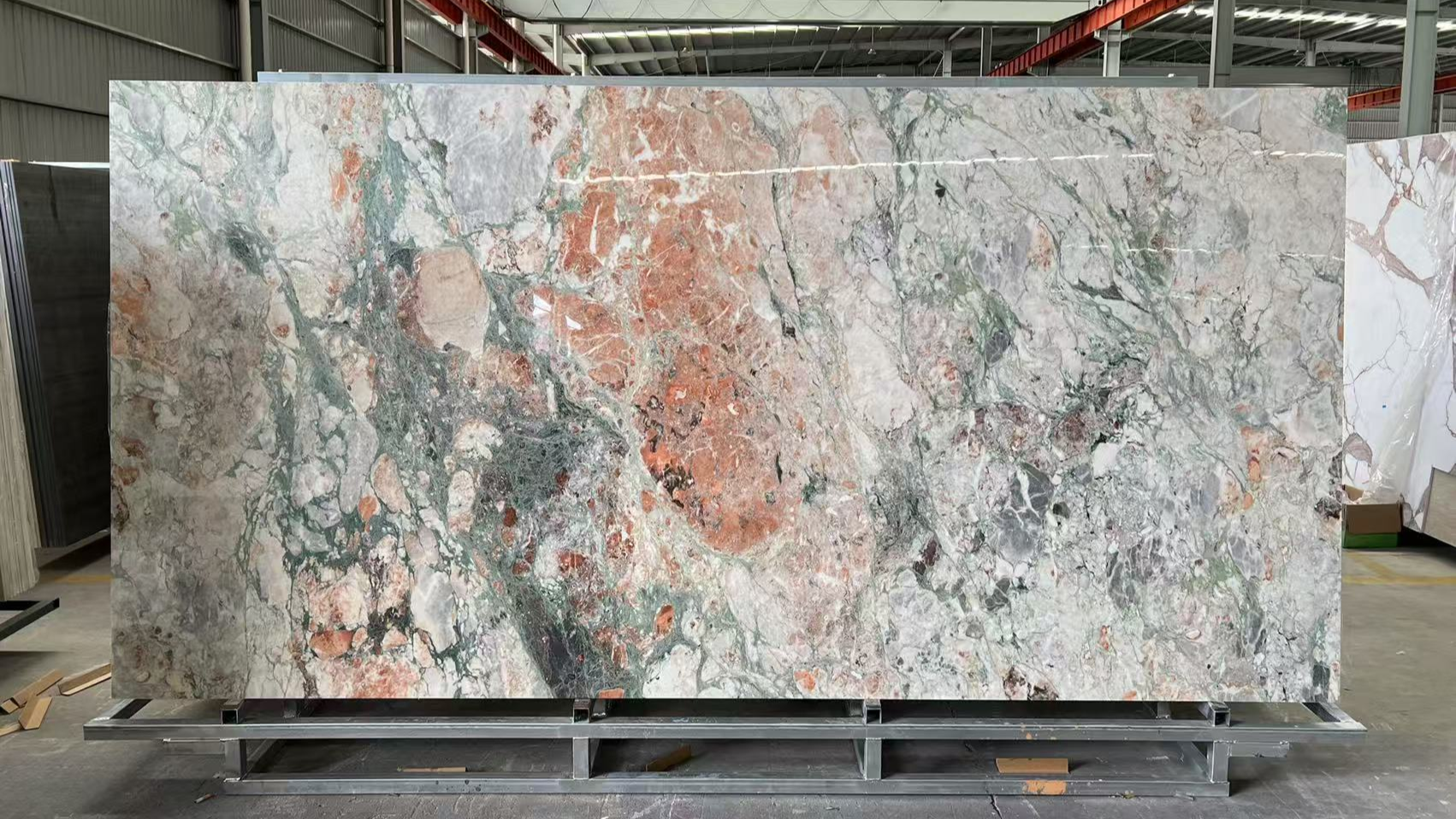 Large slab of multicolored marble with intricate green, pink, and gray veining, displayed in an industrial warehouse setting.