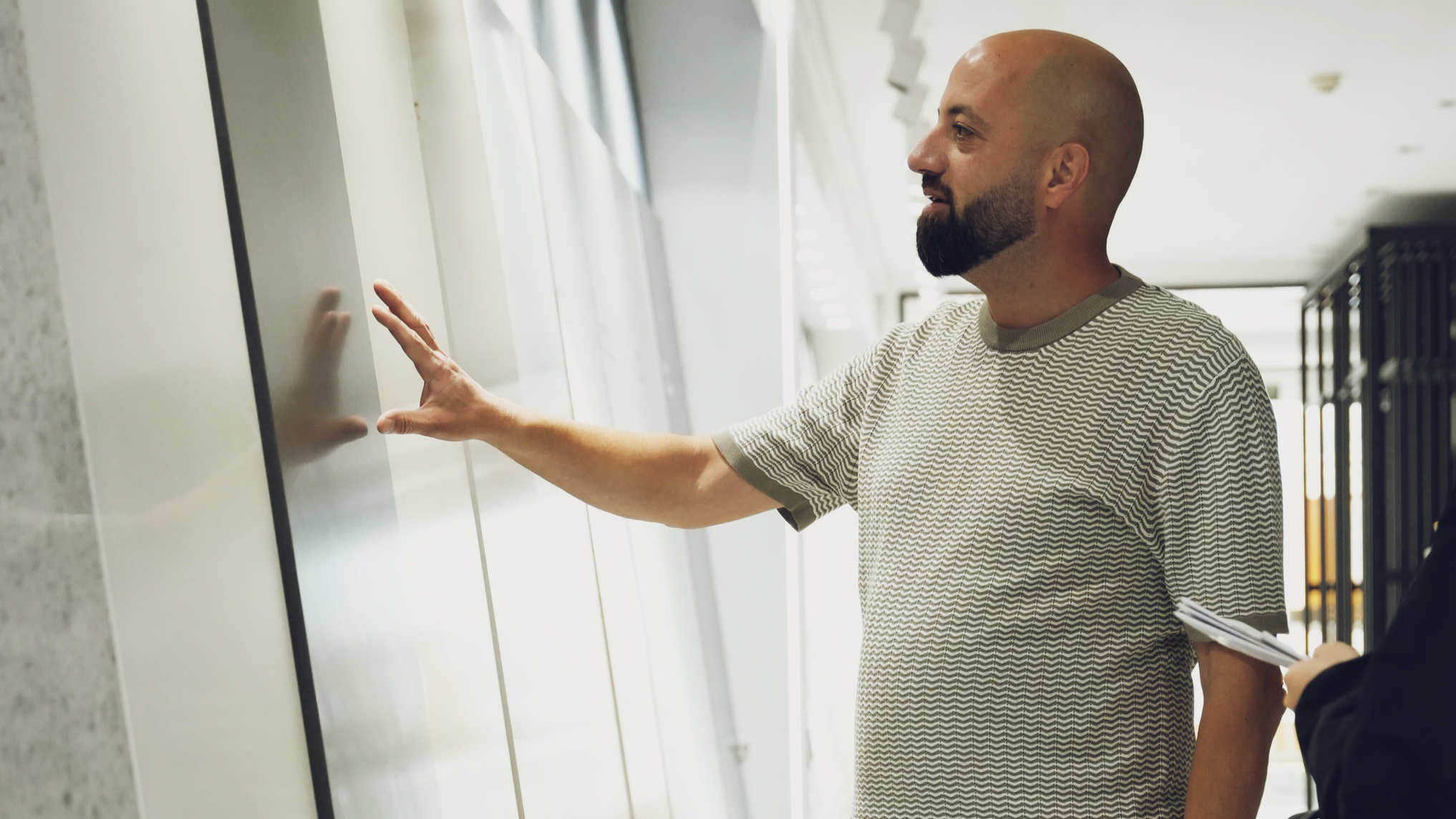 A man with a beard and bald head touching a wall-mounted whiteboard or panel, standing indoors with natural light, wearing a short-sleeved striped shirt.