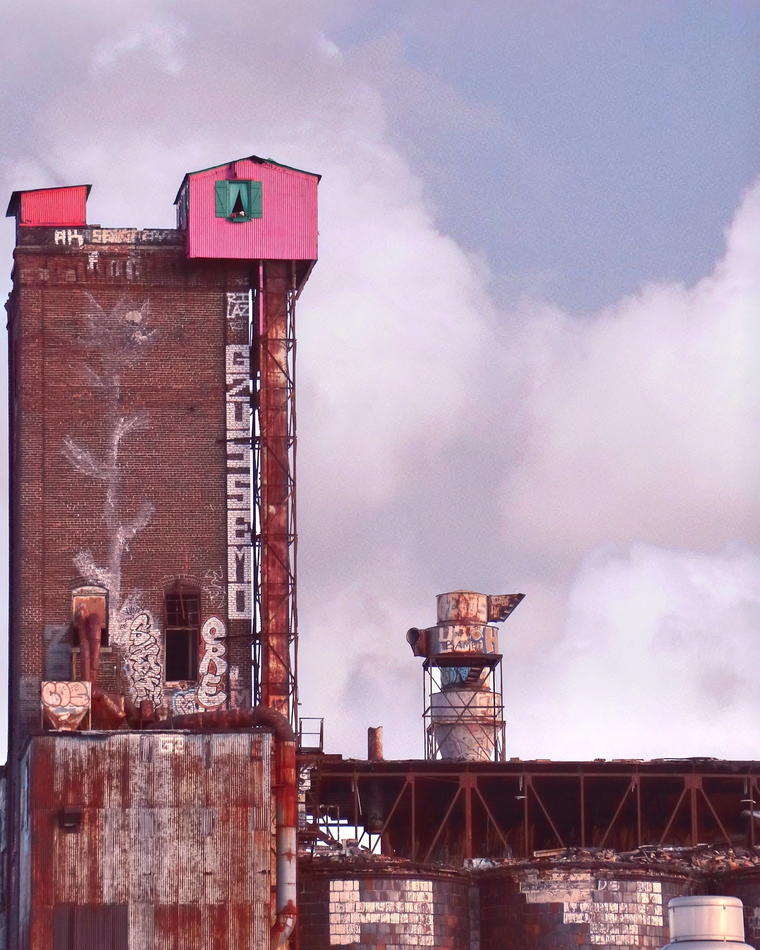 An old industrial building with graffiti on the brick wall and a pink structure on top with green shutters, rusted pipes, and metal water tanks against a cloudy sky.
