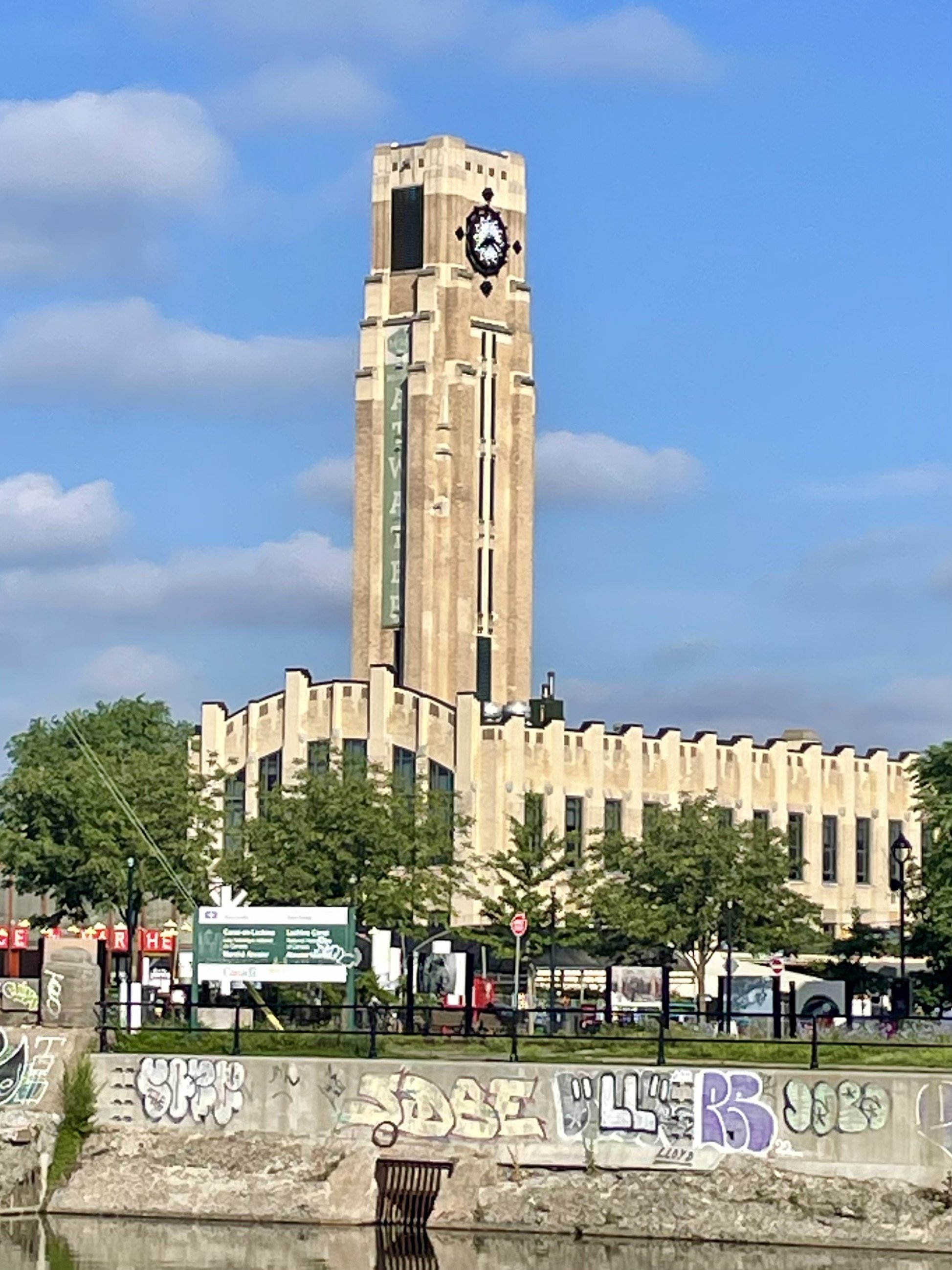 A tall clock tower with the time showing 4:15, standing behind a building with graffiti on a stone wall, with trees and a blue sky with clouds in the background.