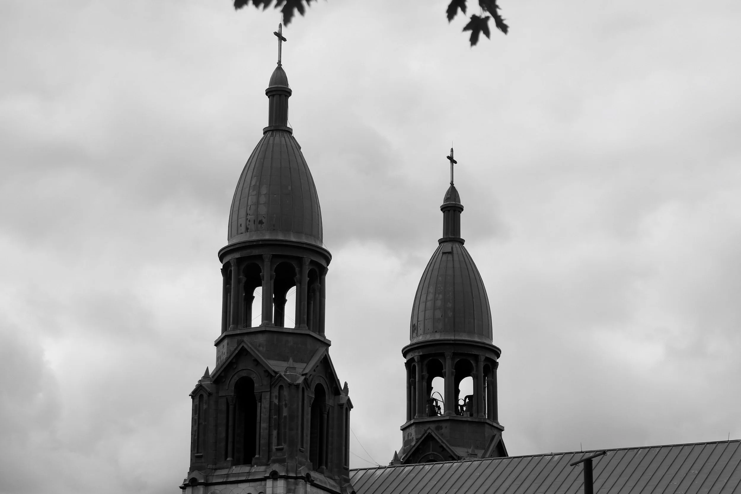 Black and white photograph of a church with two tall towers topped with crosses, against a cloudy sky, with some tree leaves visible at the top.