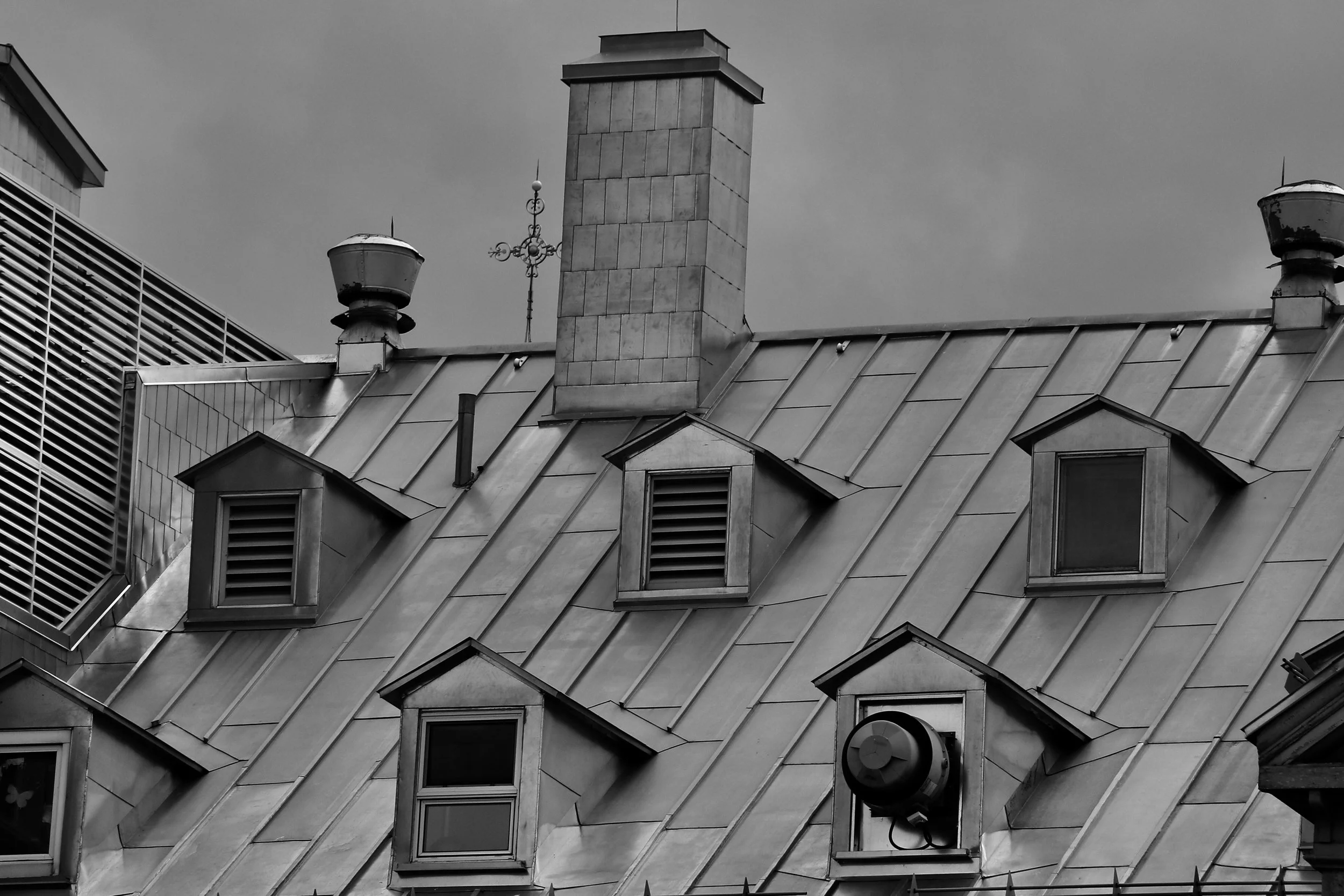 Black and white photo of a building's rooftop with multiple windows, chimneys, and vents, under a cloudy sky.