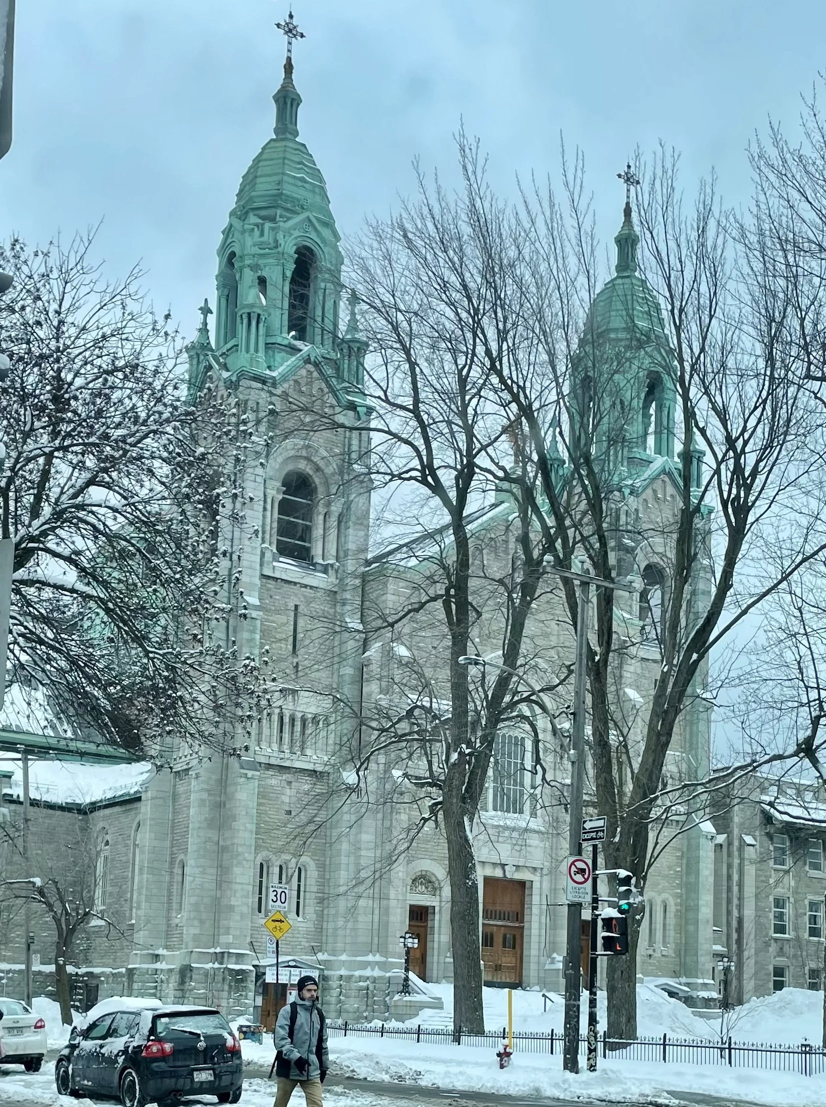 A snowy city street scene with a large historic church featuring two tall greenish spires with crosses on top. Several leafless trees and parked cars are visible, along with a pedestrian walking on the sidewalk and street signs, including a no parkin