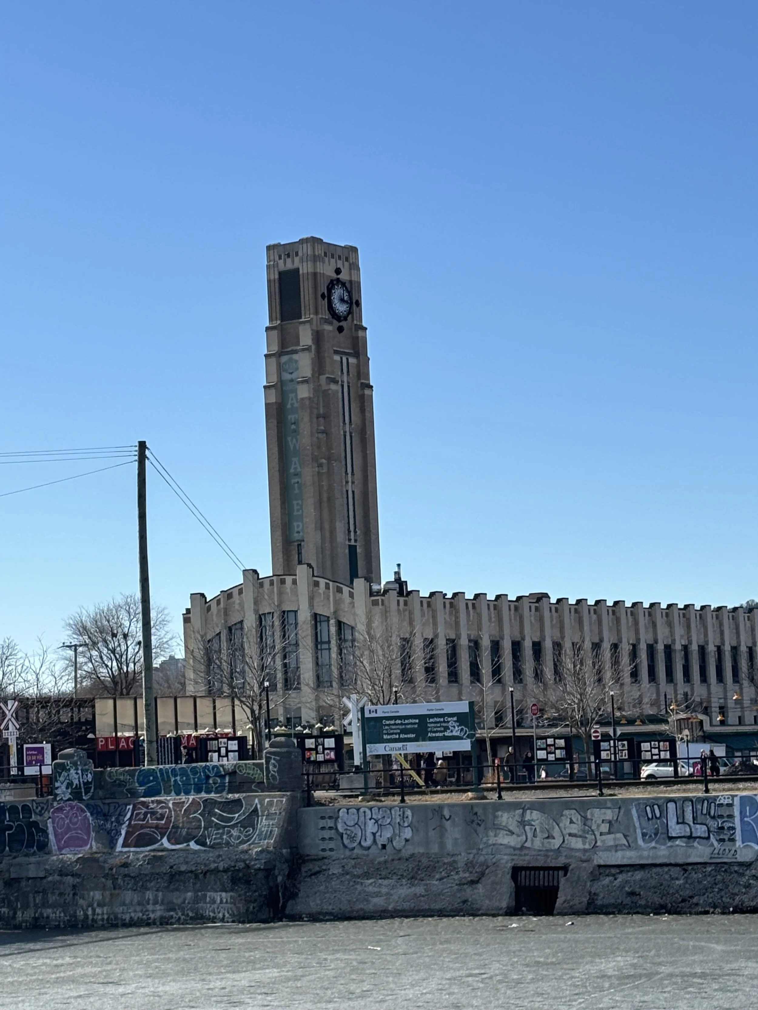 A tall clock tower with the words 'Athena' on it, under a clear blue sky. In the foreground, a concrete retaining wall covered with colorful graffiti art and street signs.