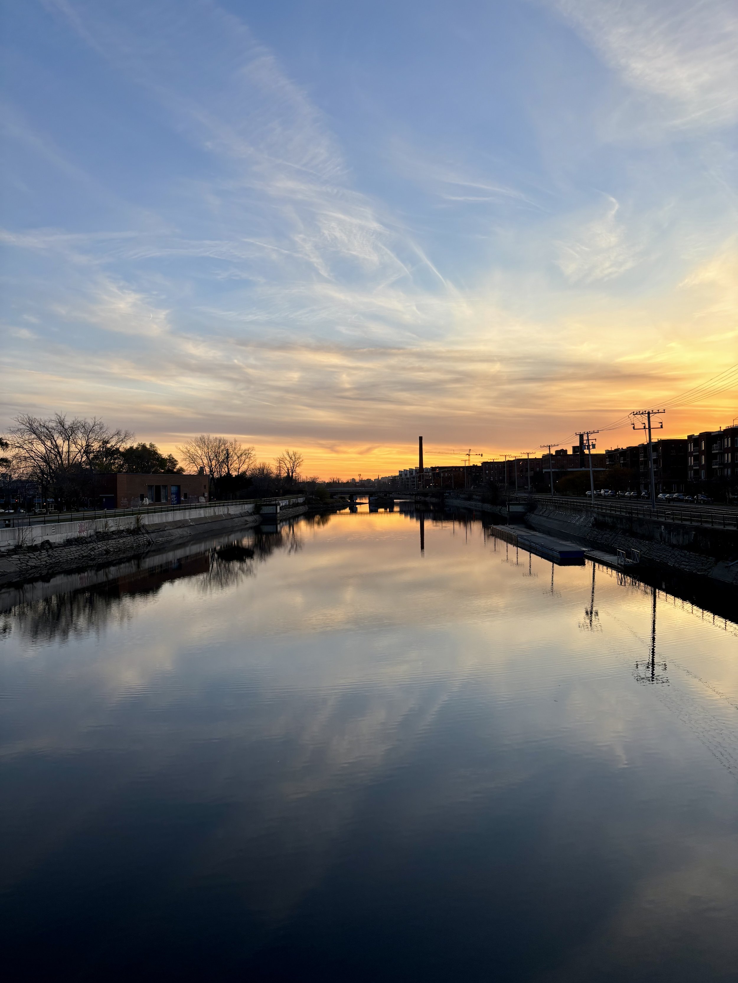 Sunset over a calm river, with a partly cloudy sky and silhouetted trees and buildings along the riverbank.