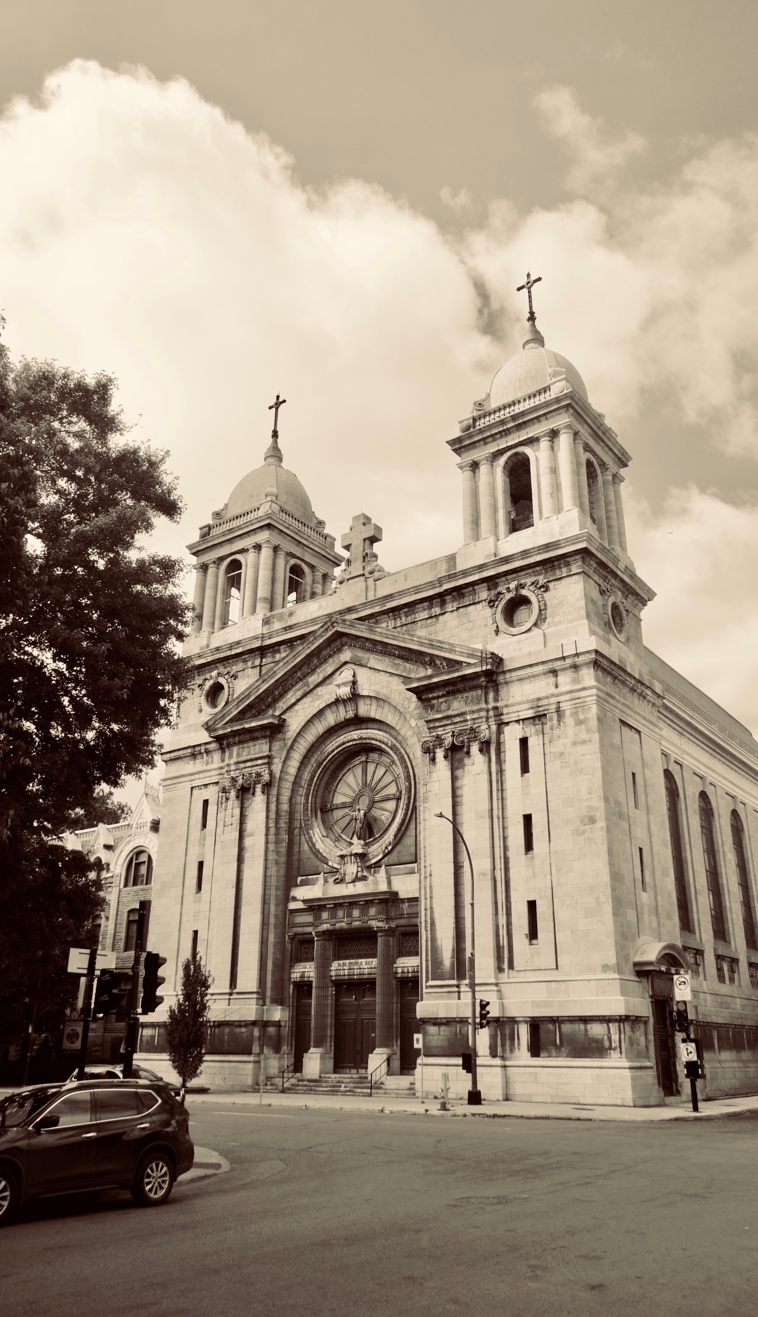 A historic church with twin towers topped by crosses, an ornate entrance, and stained glass windows, situated on a city street with cars and traffic lights.