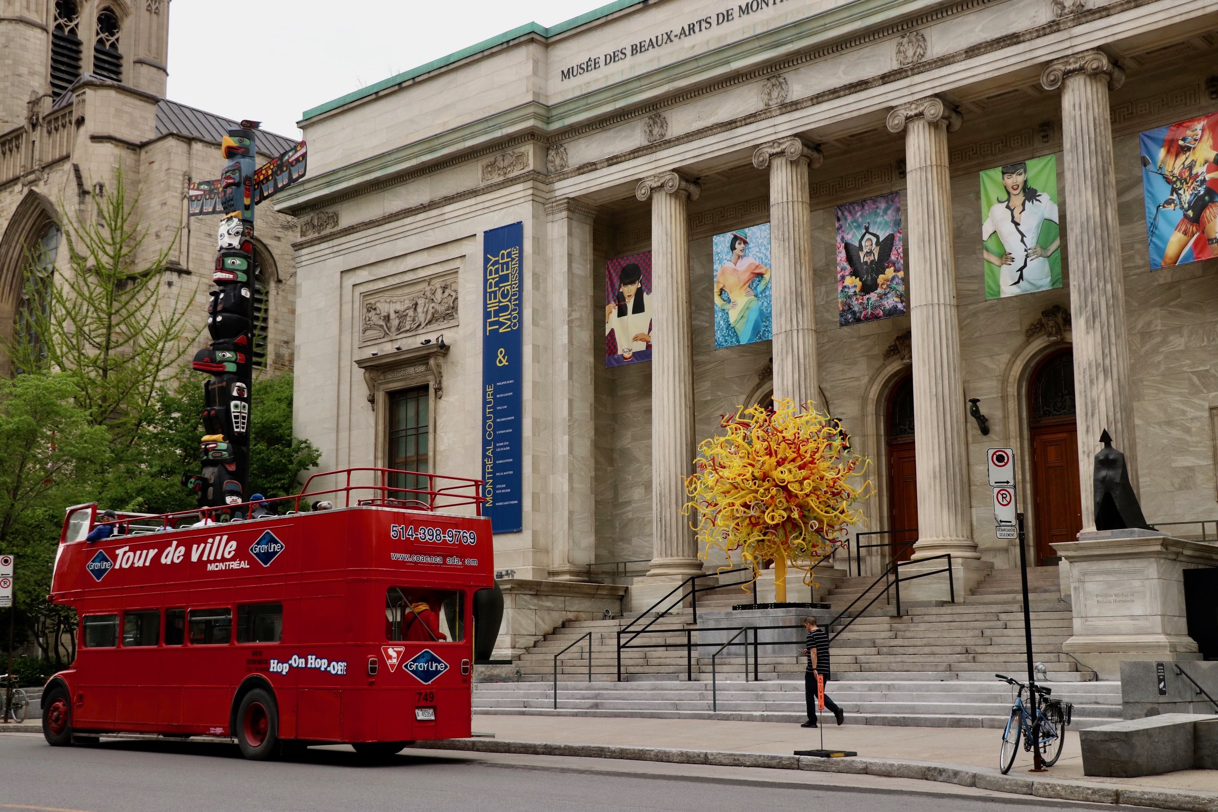 Exterior of the Montreal Museum of Fine Arts with banners displaying artwork and a large yellow glass sculpture near the entrance. A red tour bus is parked in front with a passenger on top, and a person is walking on the steps near the bus.