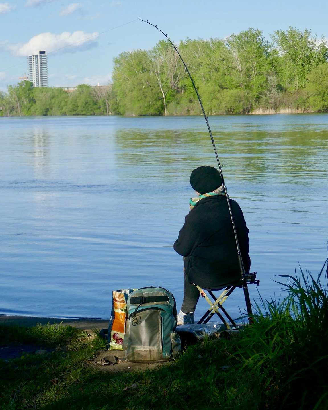 A person fishing by a riverbank, sitting on a small chair with a fishing rod cast into the water. There is a backpack and a bag on the ground nearby, with trees and buildings visible in the background.