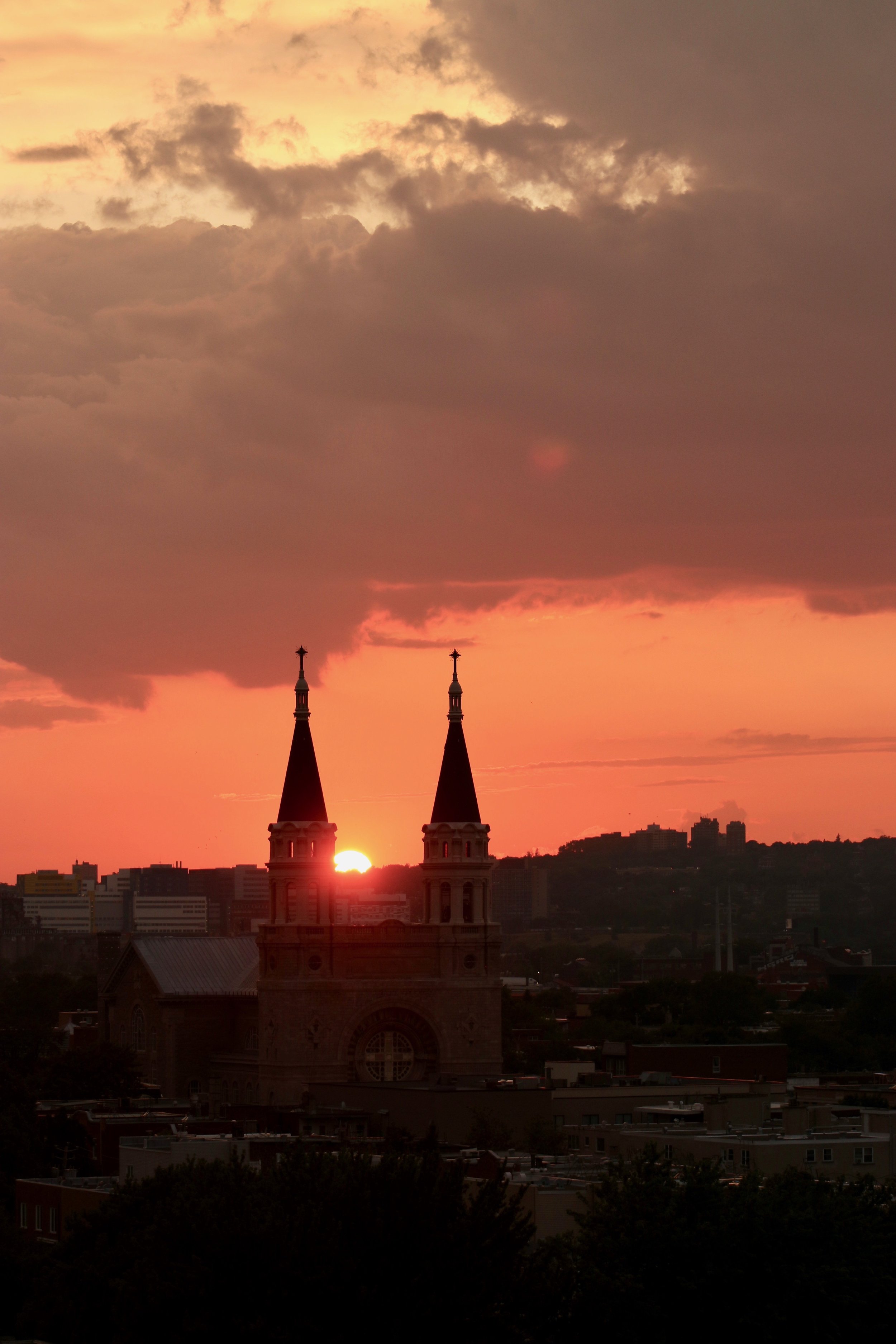Sunset over a city skyline with two church steeples in the foreground and clouds in the sky.