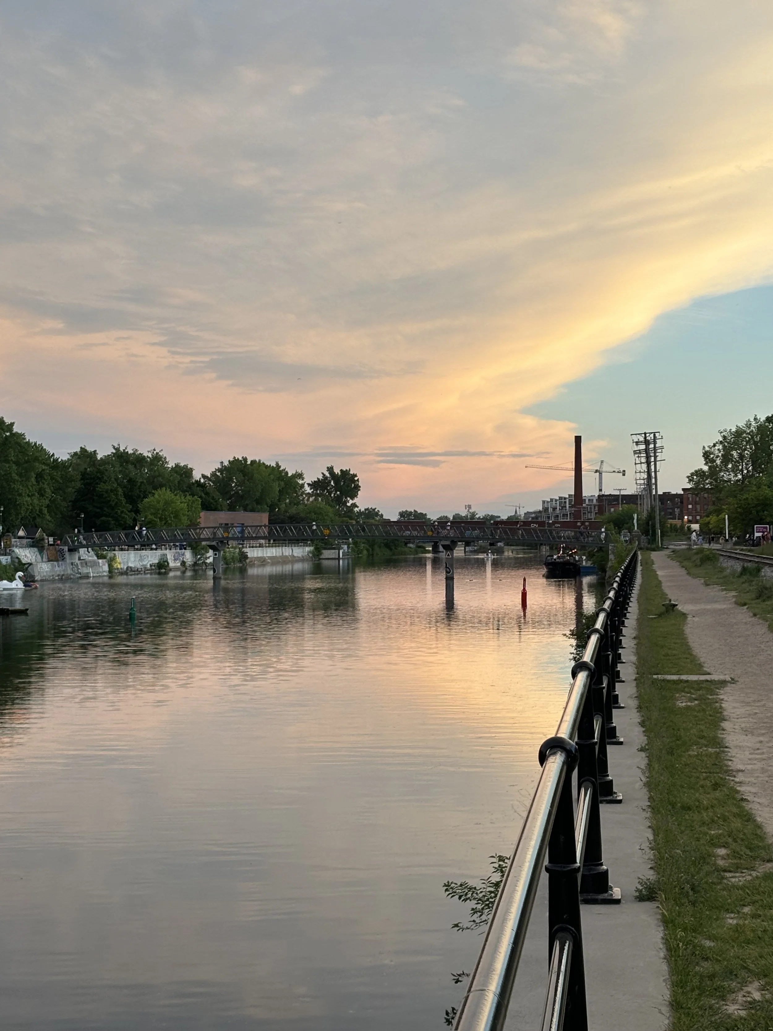 A peaceful riverside scene during sunset with a bridge over the water, trees lining the riverbank, a walkway with a metal railing, and a distant old factory with a chimney and cranes in the background.