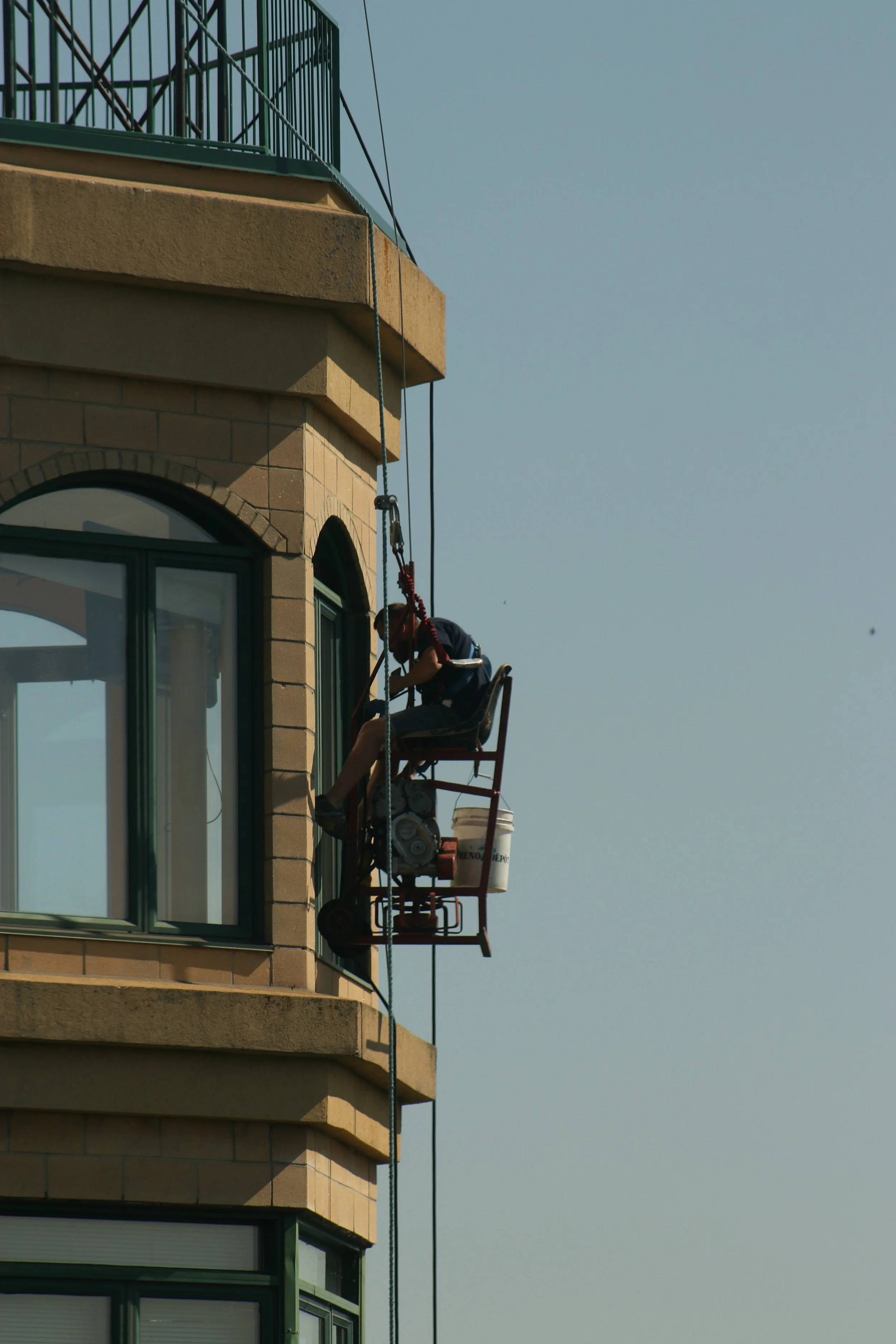 A worker on a suspended platform cleaning or inspecting the exterior of a tall building with multiple windows and balconies against a clear sky.