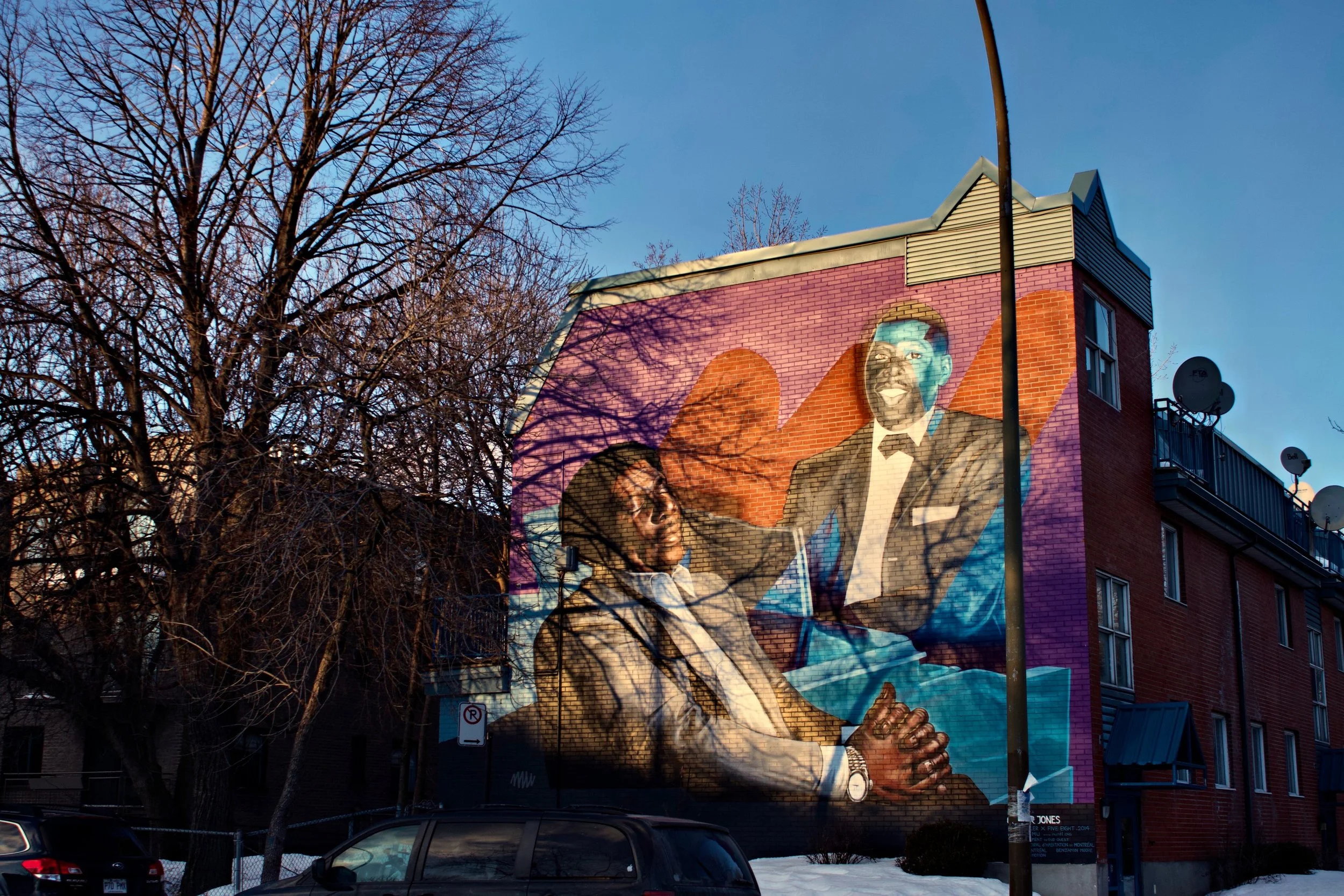 Colorful mural of two men in suits shaking hands and smiling on the side of a red brick building, with leafless trees and a blue sky in the background.