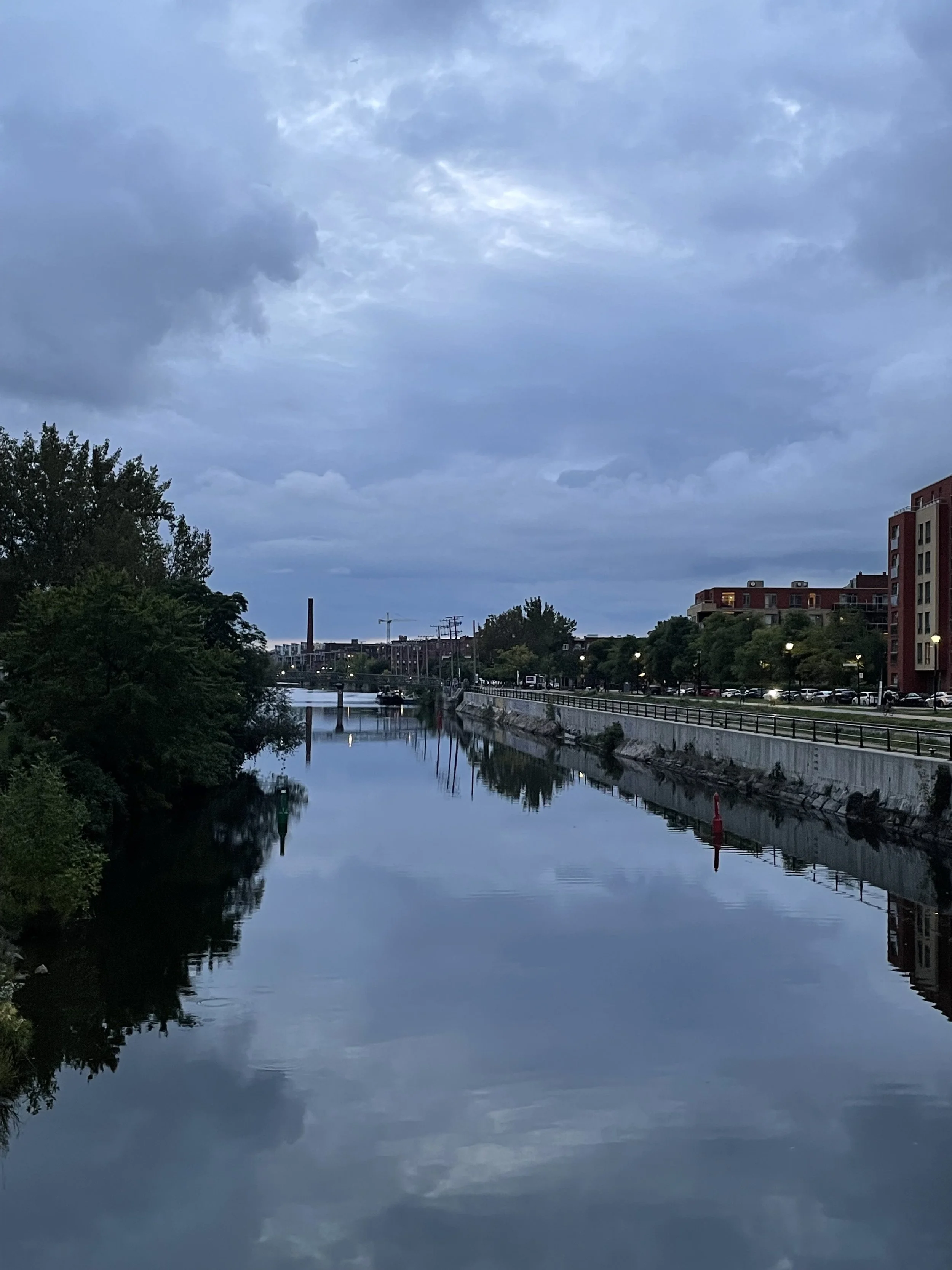 Overcast sky above a calm city canal with trees on the left and a brick building on the right, reflections of the sky and buildings visible in the water.