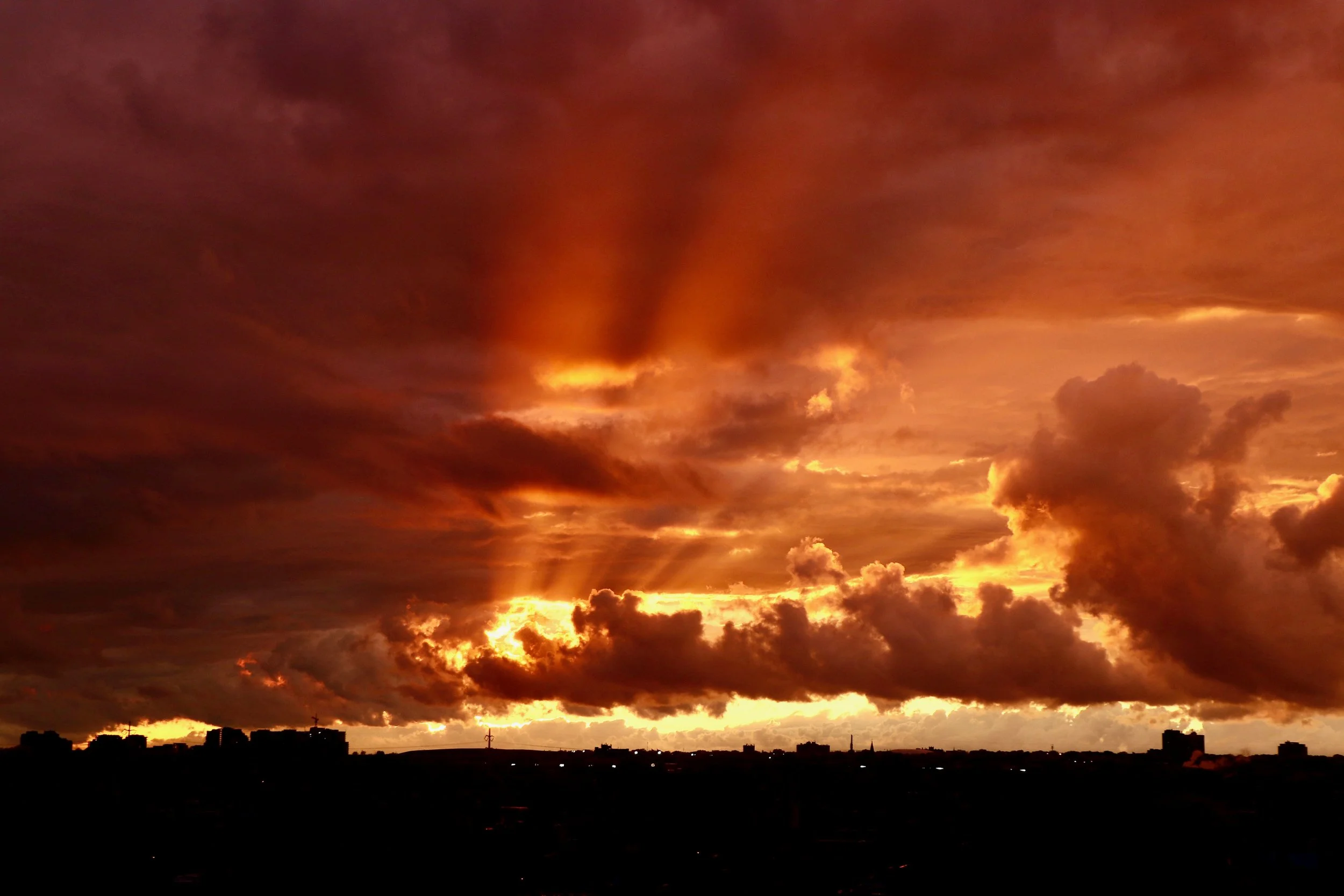 A dramatic sky filled with dark clouds illuminated by the orange and yellow hues of a setting or rising sun, with silhouettes of buildings and power lines on the horizon.