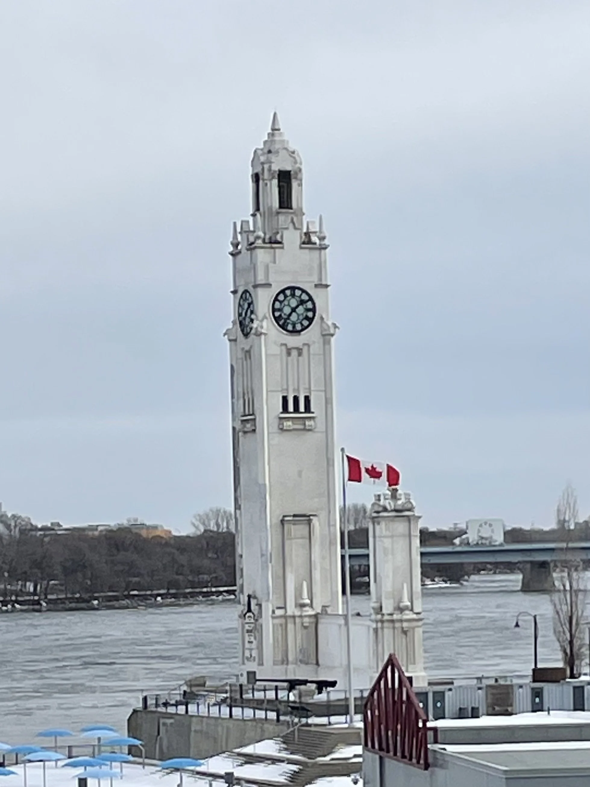 Clock tower with a Canadian flag near a waterfront with docked boats and umbrellas.