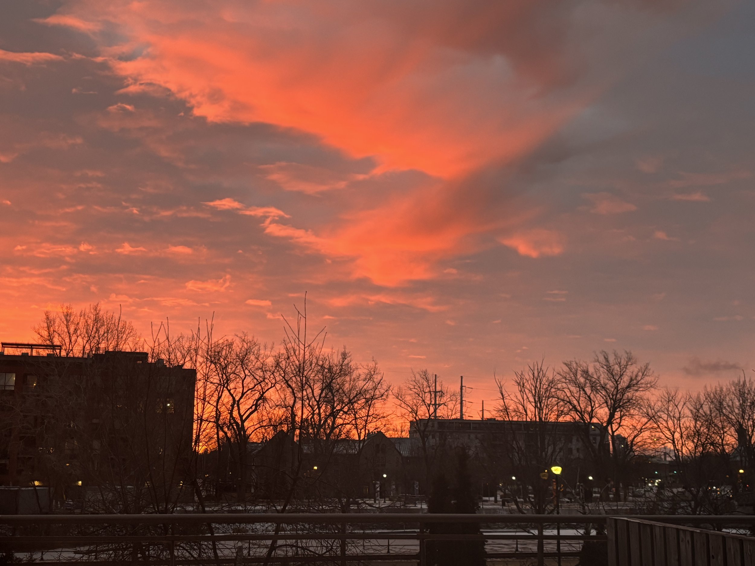 A sunset sky with pink and orange clouds over a silhouetted cityscape with trees and buildings.
