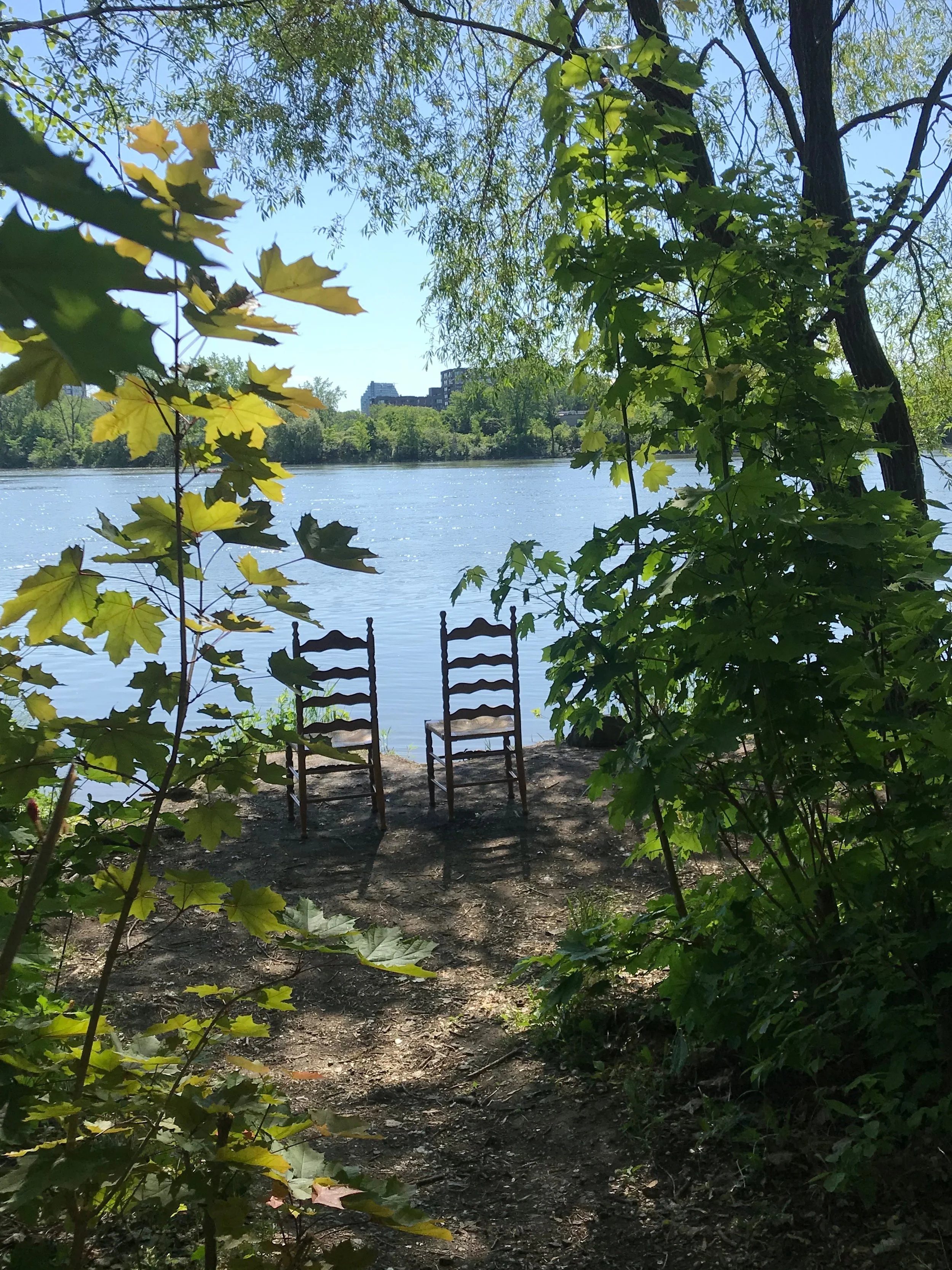 Two chairs facing a river, surrounded by green trees and foliage, with buildings in the background on a sunny day.