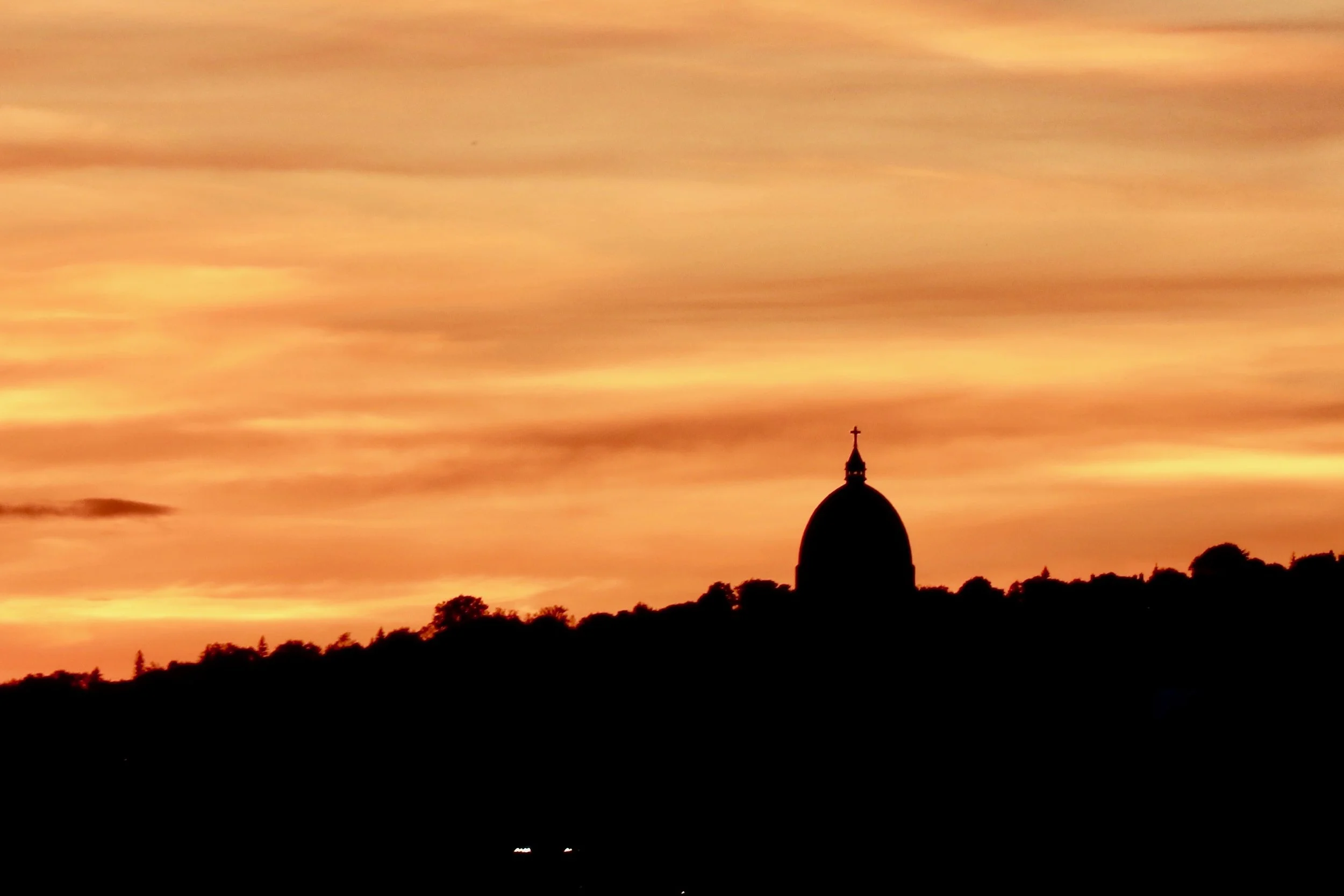 Sunset sky with orange and pink clouds, silhouette of a church steeple with a cross, and tree line in the foreground.