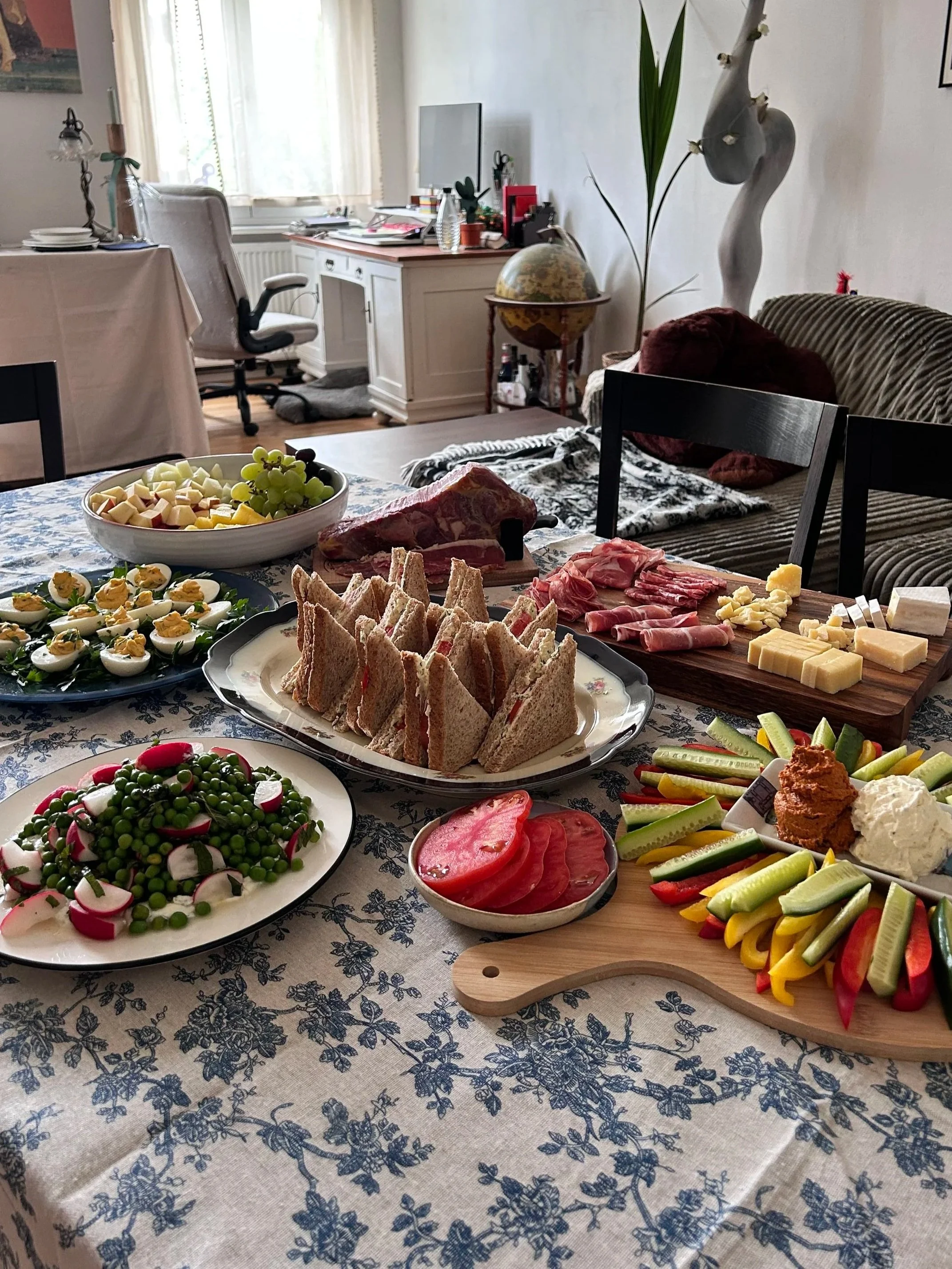 A dining table with an assortment of foods including sandwiches, deviled eggs, sliced meats, cheese, vegetables, and a fruit platter, set on a tablecloth with a blue floral pattern, in a room with a sideboard, a globe, and decorative items.