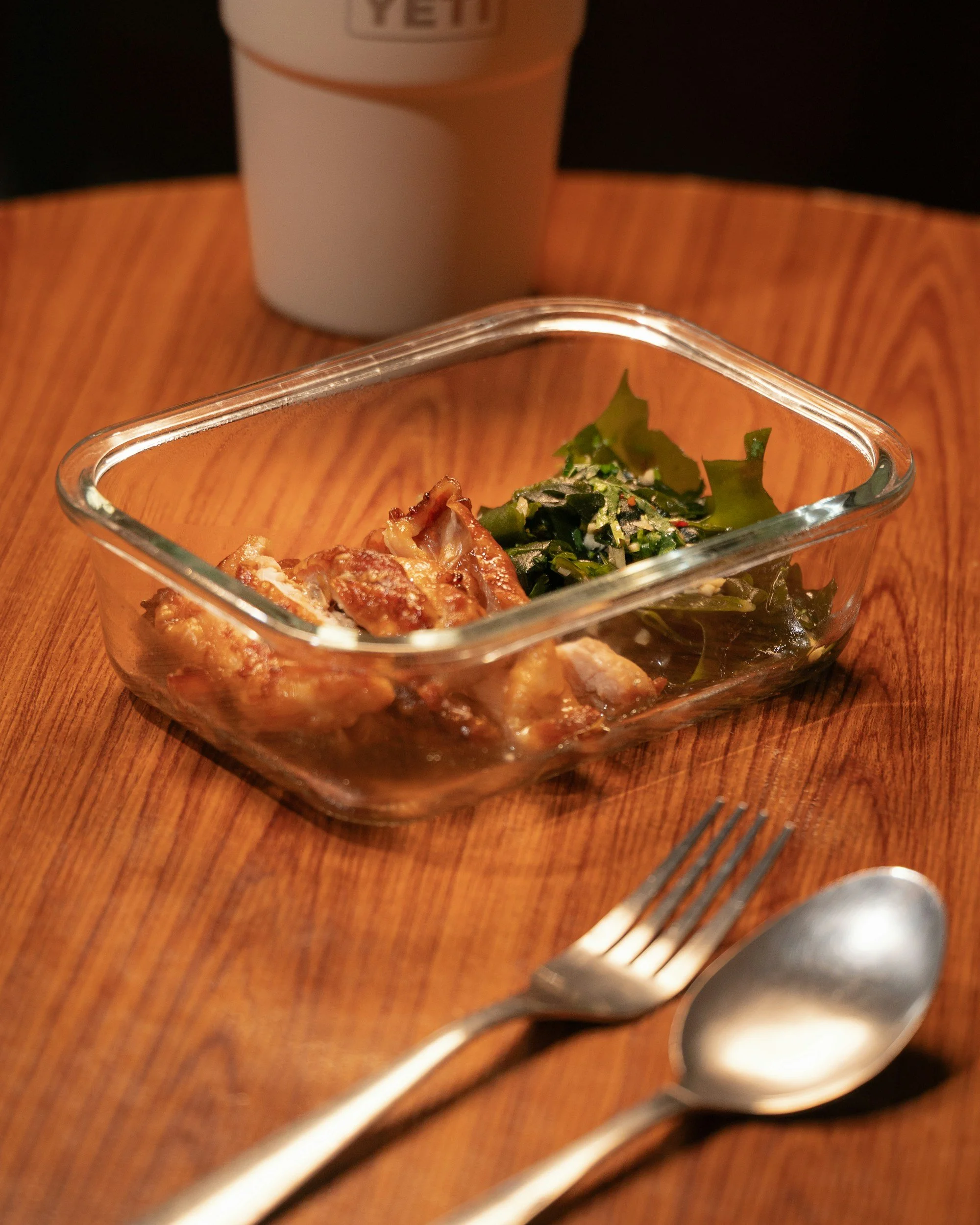 A glass dish of fried chicken and a side salad with greens on a wooden table. A fork and spoon are in the foreground, and a white beverage cup is in the background.