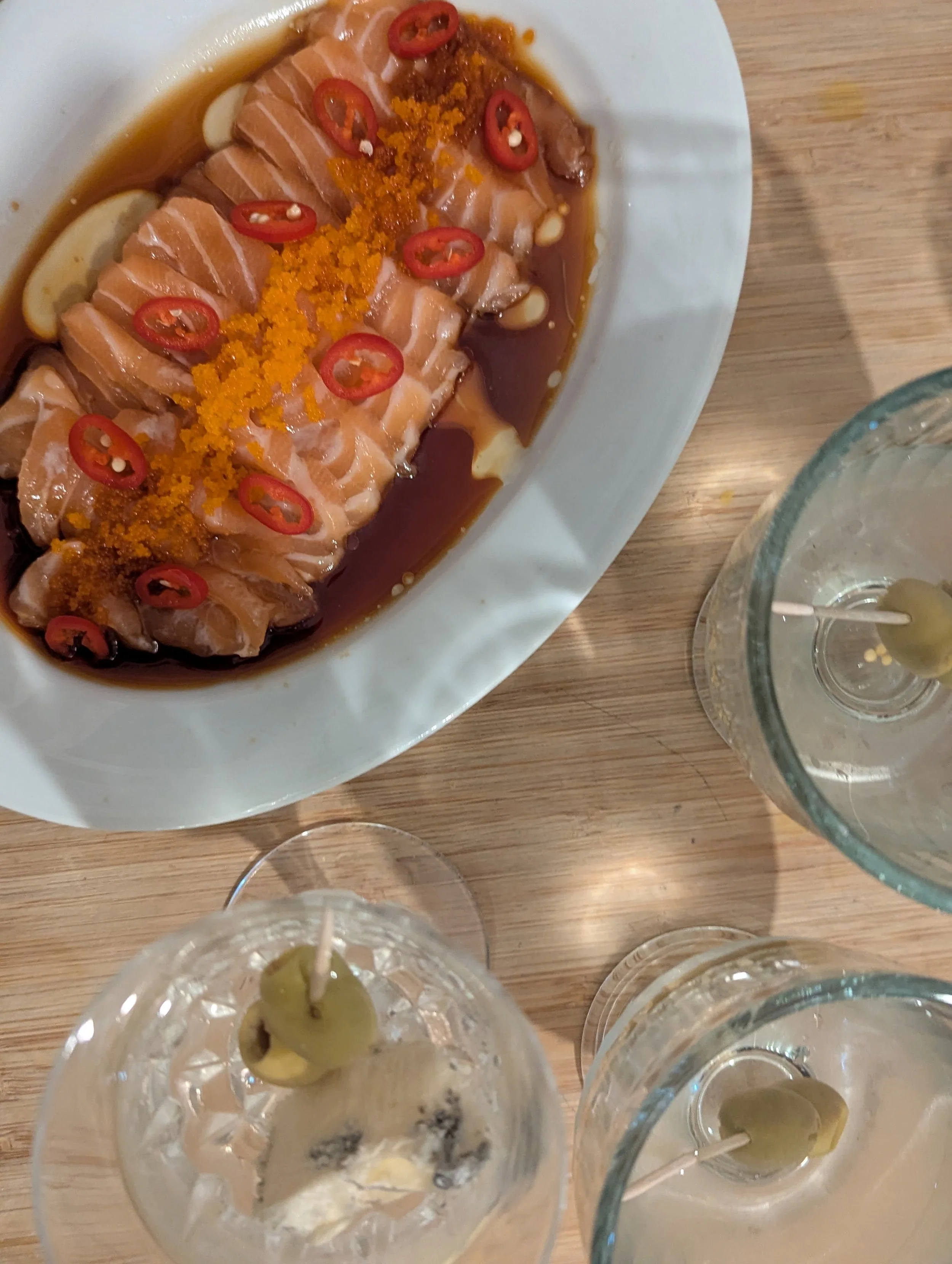 A white oval plate with slices of raw salmon topped with sliced red chili peppers, orange seasoning, and surrounded by sauce, placed on a wooden table. Several glasses with appetizers are partially visible around the main plate.