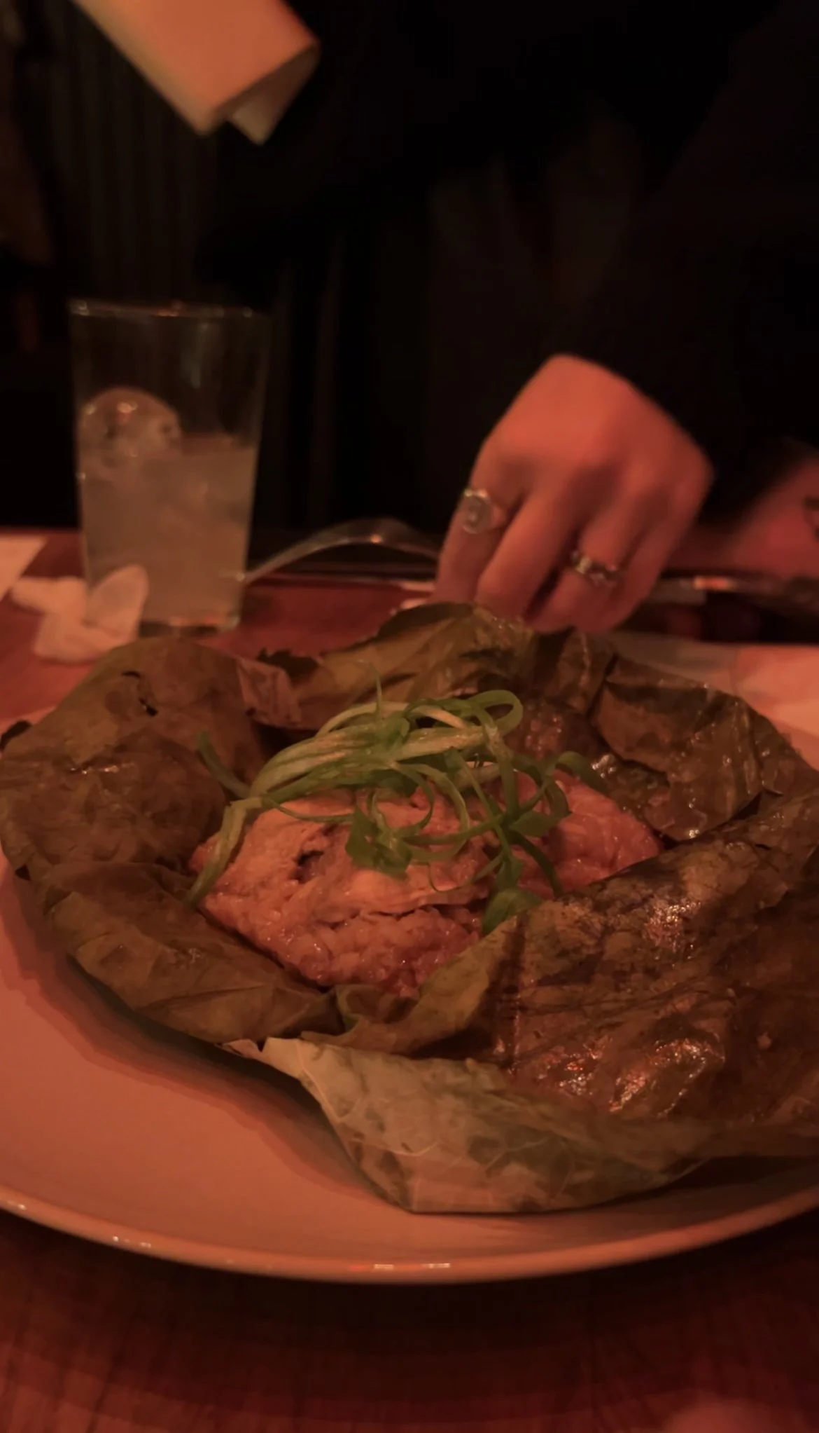 A person is serving food from a large tray lined with leaf, containing grilled meats and garnished with green herbs, with a glass of water nearby in a dimly lit setting.