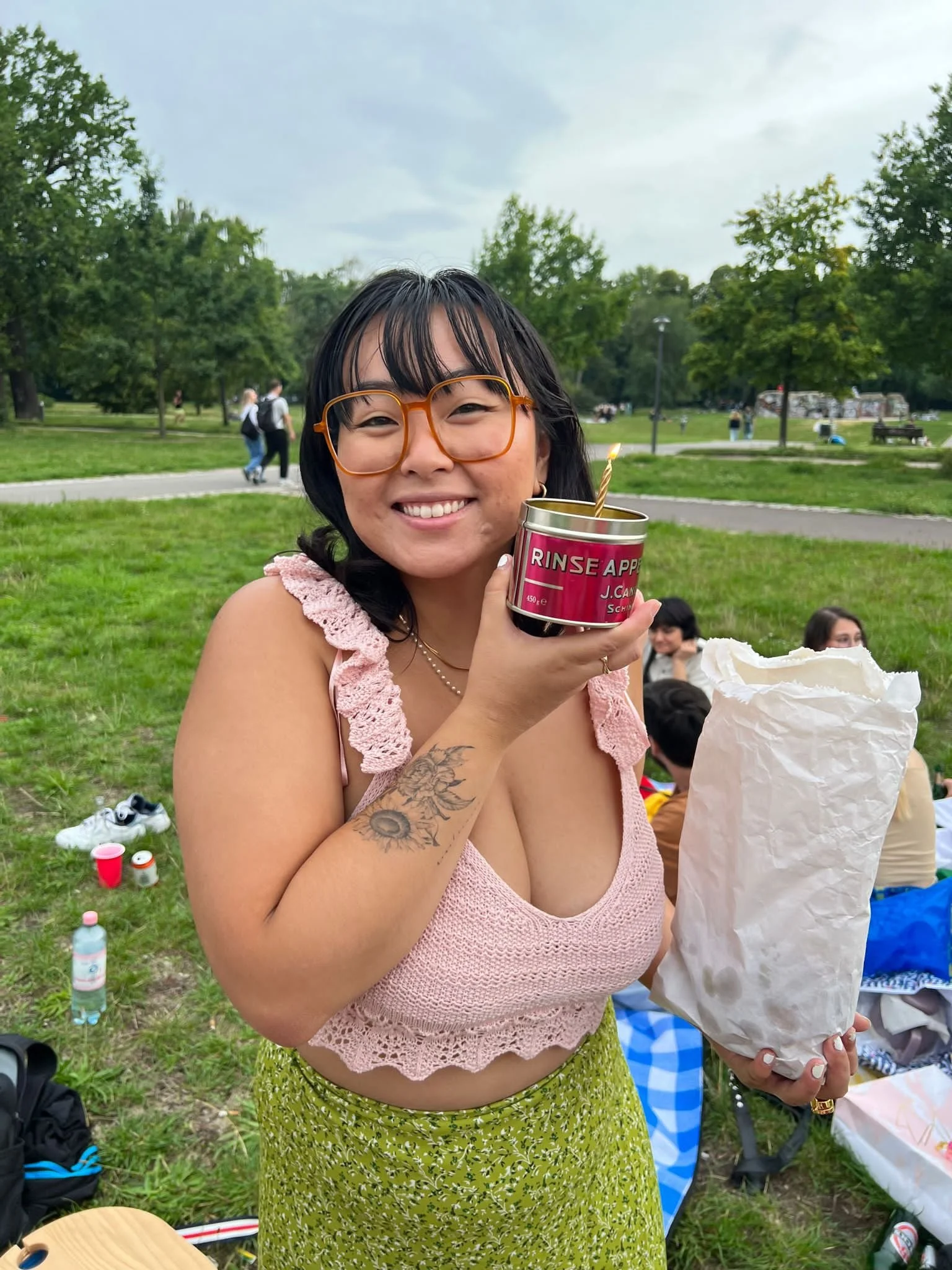 A smiling woman with black hair and glasses holding a can with a candle inside at a park during daytime. She has tattoos on her left arm and is wearing a pink lace top and a green skirt. People and trees are visible in the background.