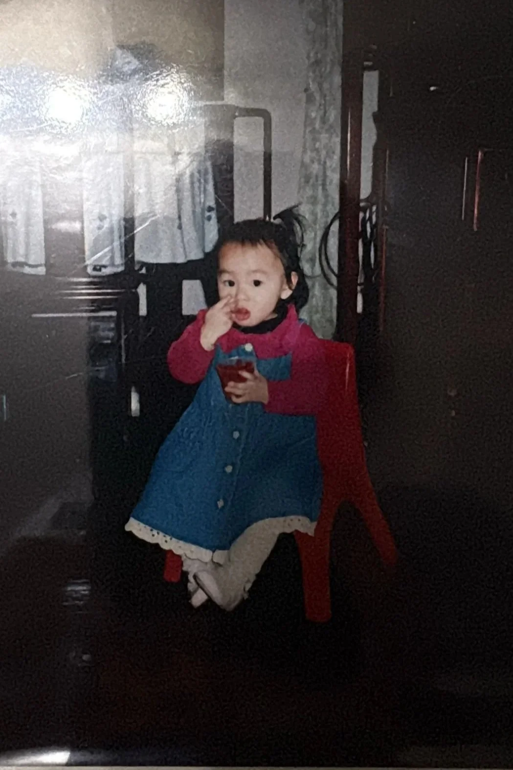 A young child with black hair in a red, blue, and white dress sitting on a red chair, holding a cup, and touching her nose with her finger. The background shows a wall and some industrial equipment.