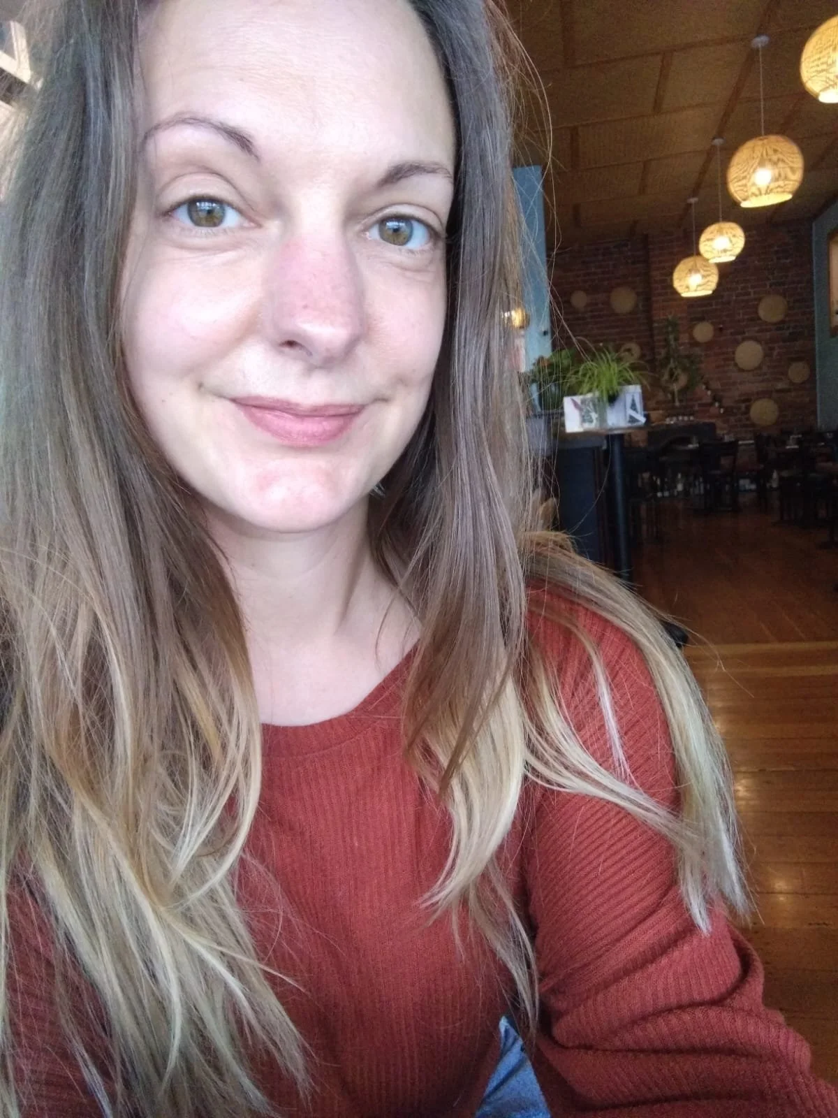 A woman with long, wavy, blonde hair and fair skin smiling while sitting inside a cozy restaurant with hanging lights and brick walls.